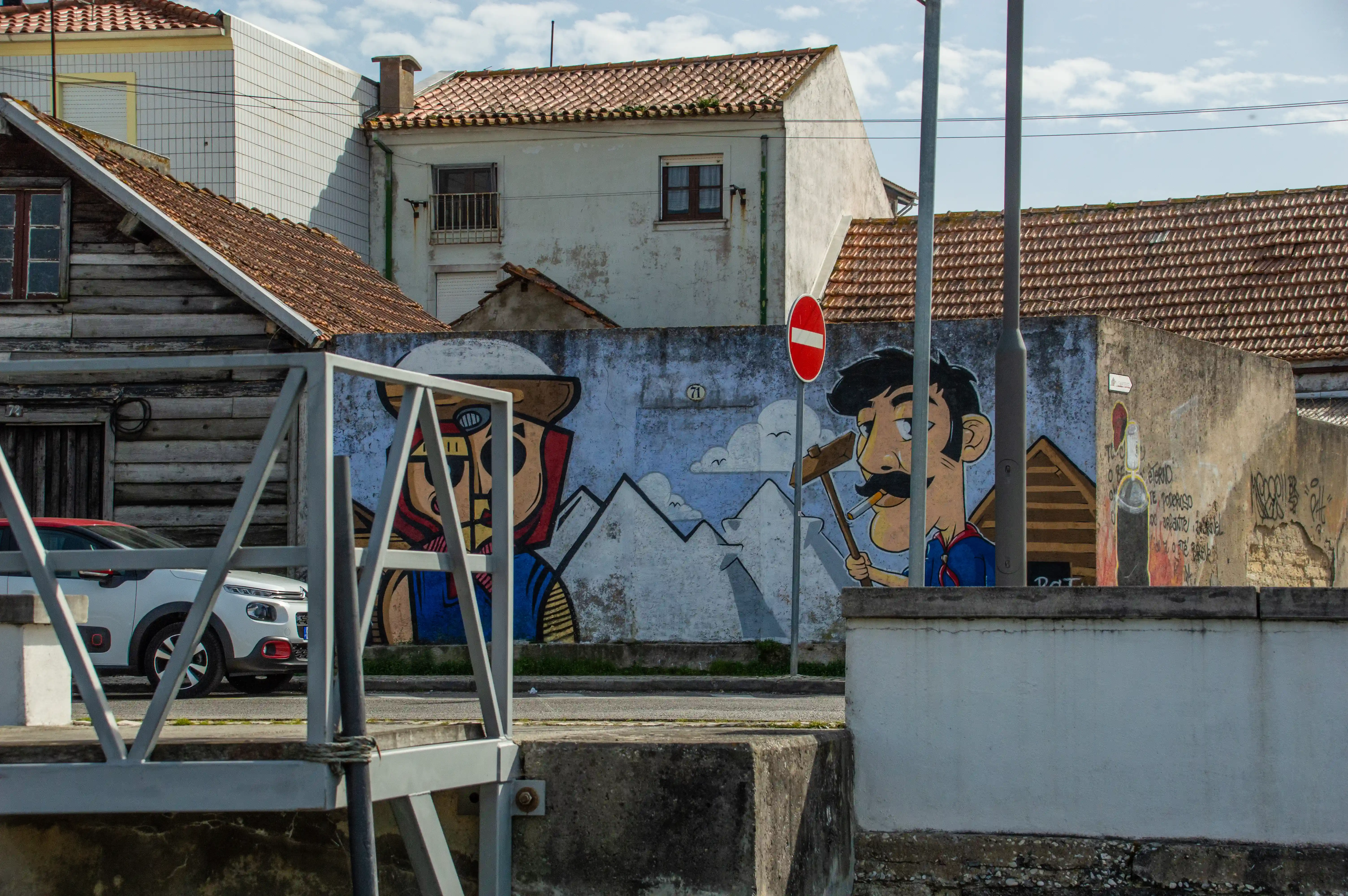 A colourful mural on a blue wall shows two cartoon characters, one with a hammer and cigarette, set against a backdrop of white mountains and clouds. Traditional buildings and a red 'No Entry' sign are also visible.