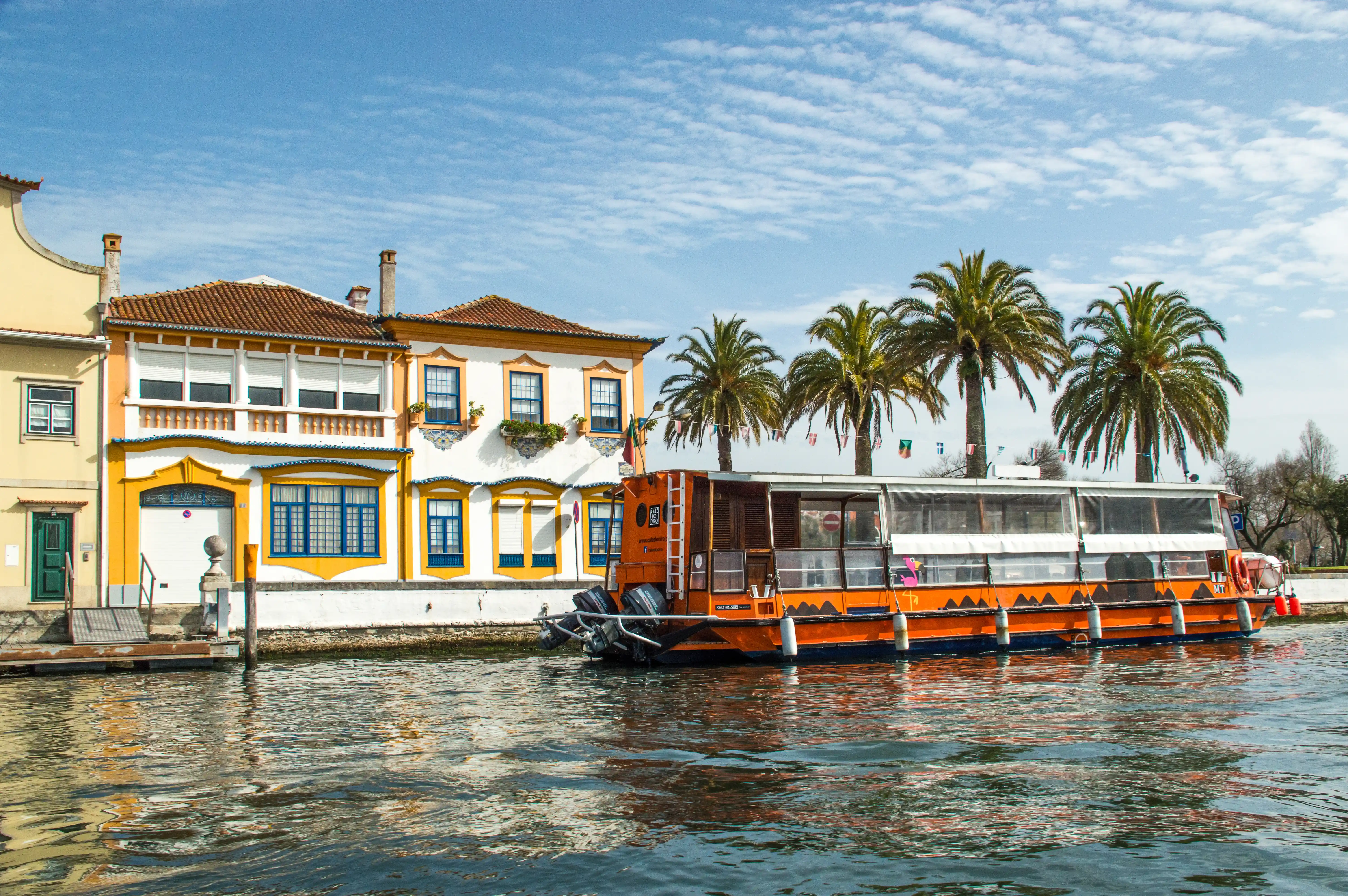 A side view of an orange tourist boat docked beside colourful traditional Portuguese buildings with palm trees in the background, reflecting on the water under a blue sky with clouds.