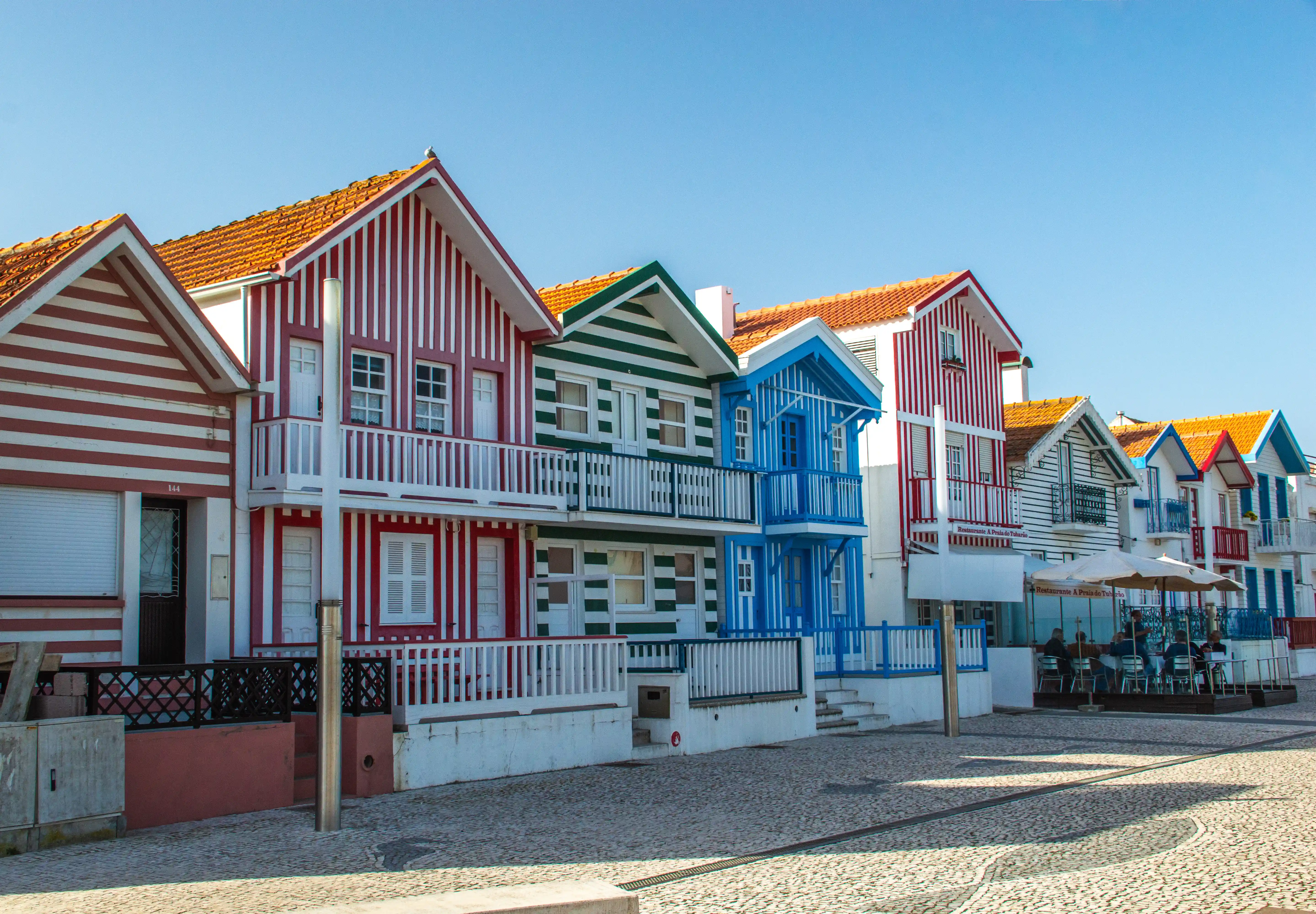 A line of charming, candy-striped houses with red, green, and blue patterns, featuring white balconies and orange roofs, are set against a bright blue sky in a coastal town.