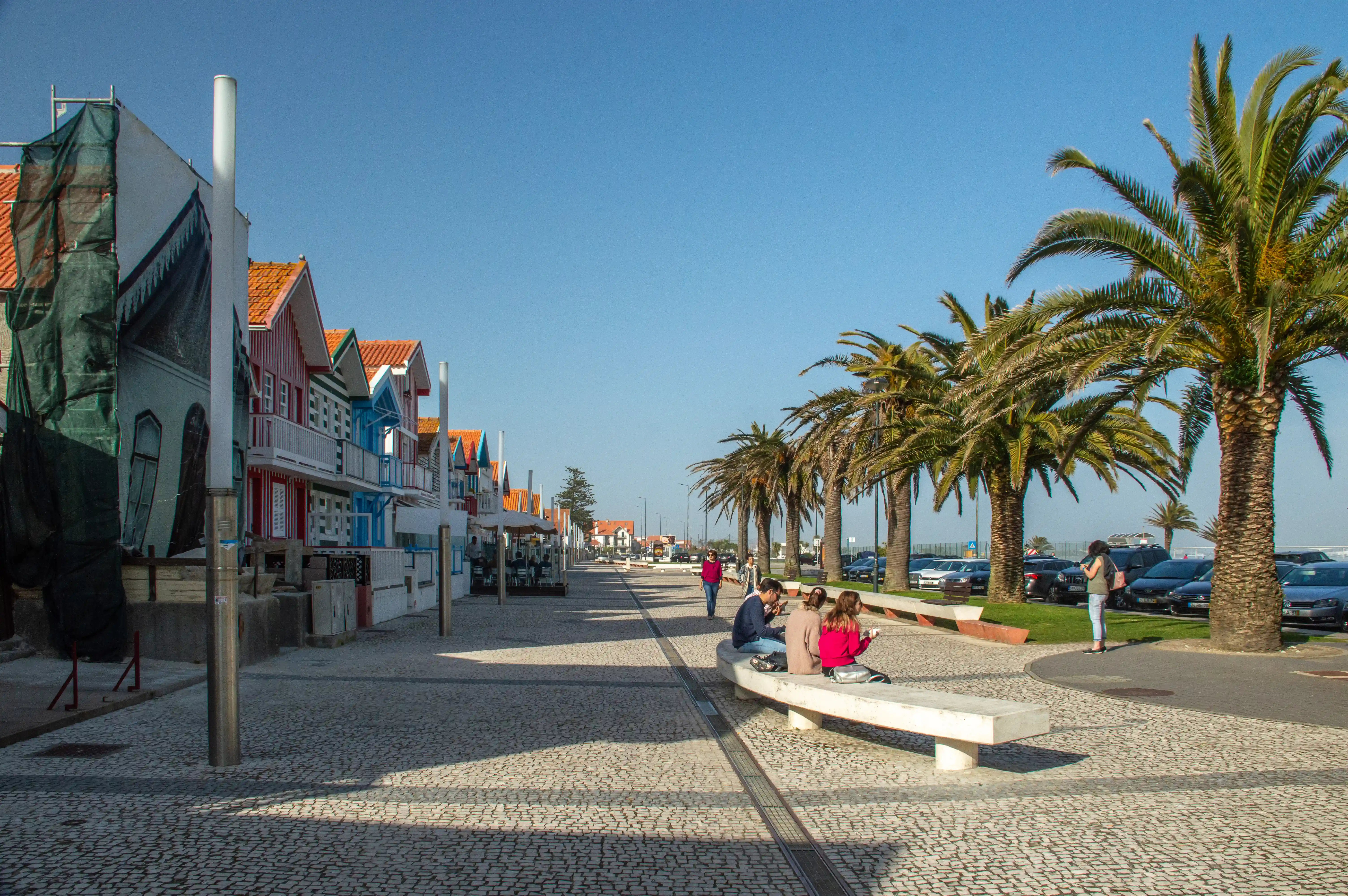 A sunny promenade in front of colourful beach houses, with palm trees and people relaxing on a bench.