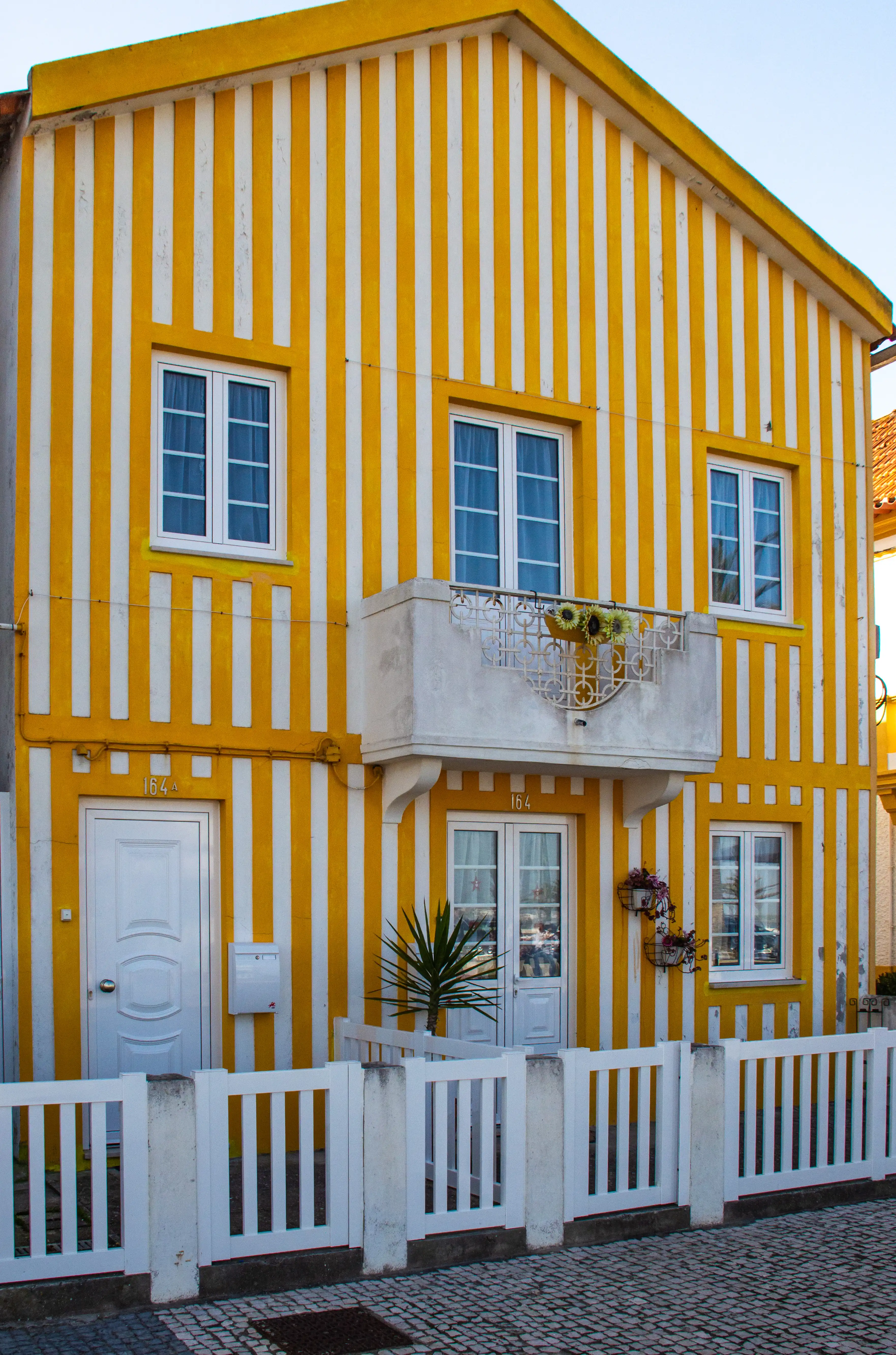 A narrow, tall house painted with bold vertical stripes of yellow and white. The house has white-framed windows, a decorative white balcony on the first floor, a white front door, and a white picket fence in front.