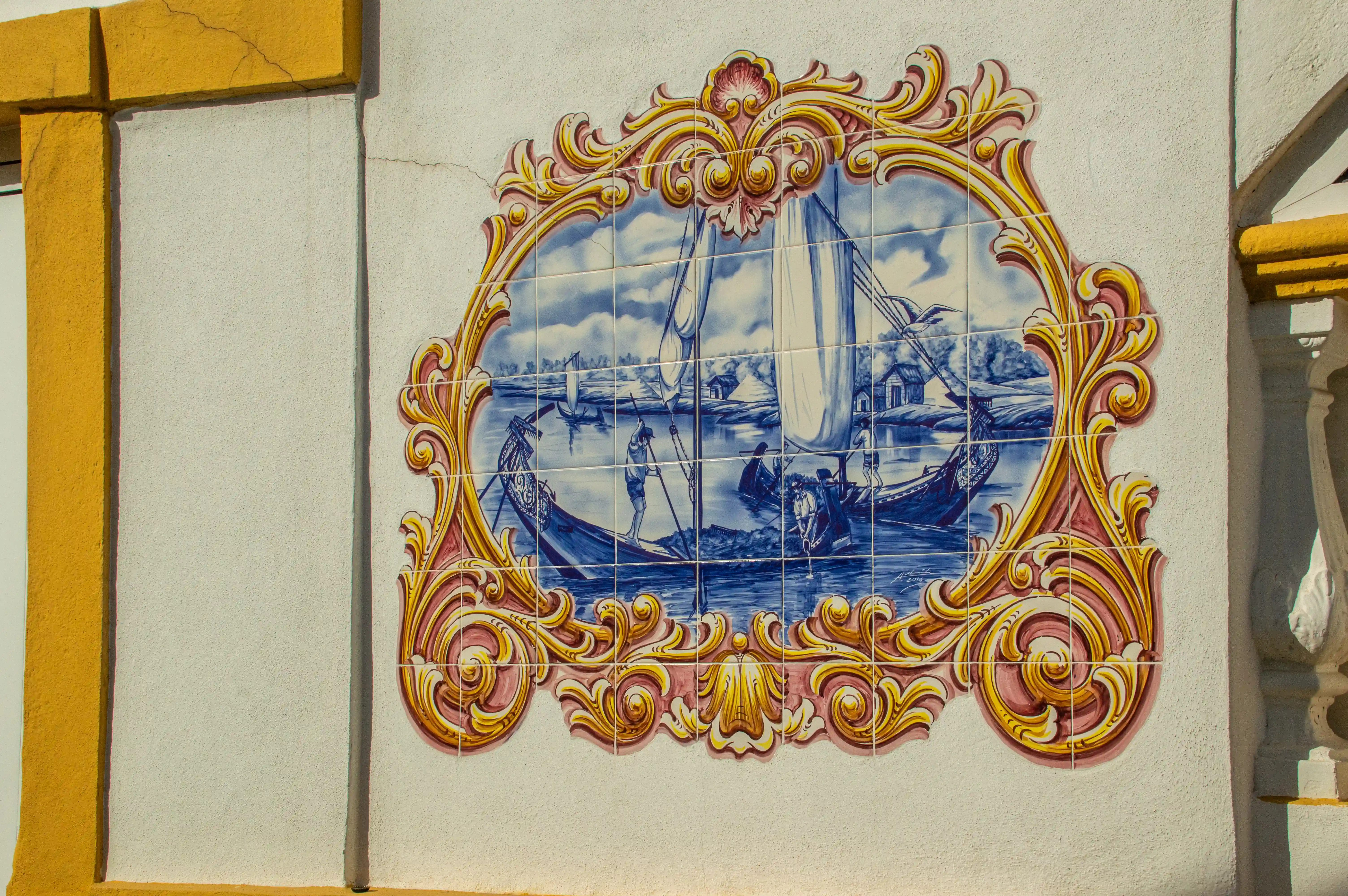 A close-up view of a decorative tiled mural on a white wall. The mural is framed by elaborate gold scrollwork and features a blue and white depiction of boats on water, with figures and distant buildings. Yellow trim borders the wall.