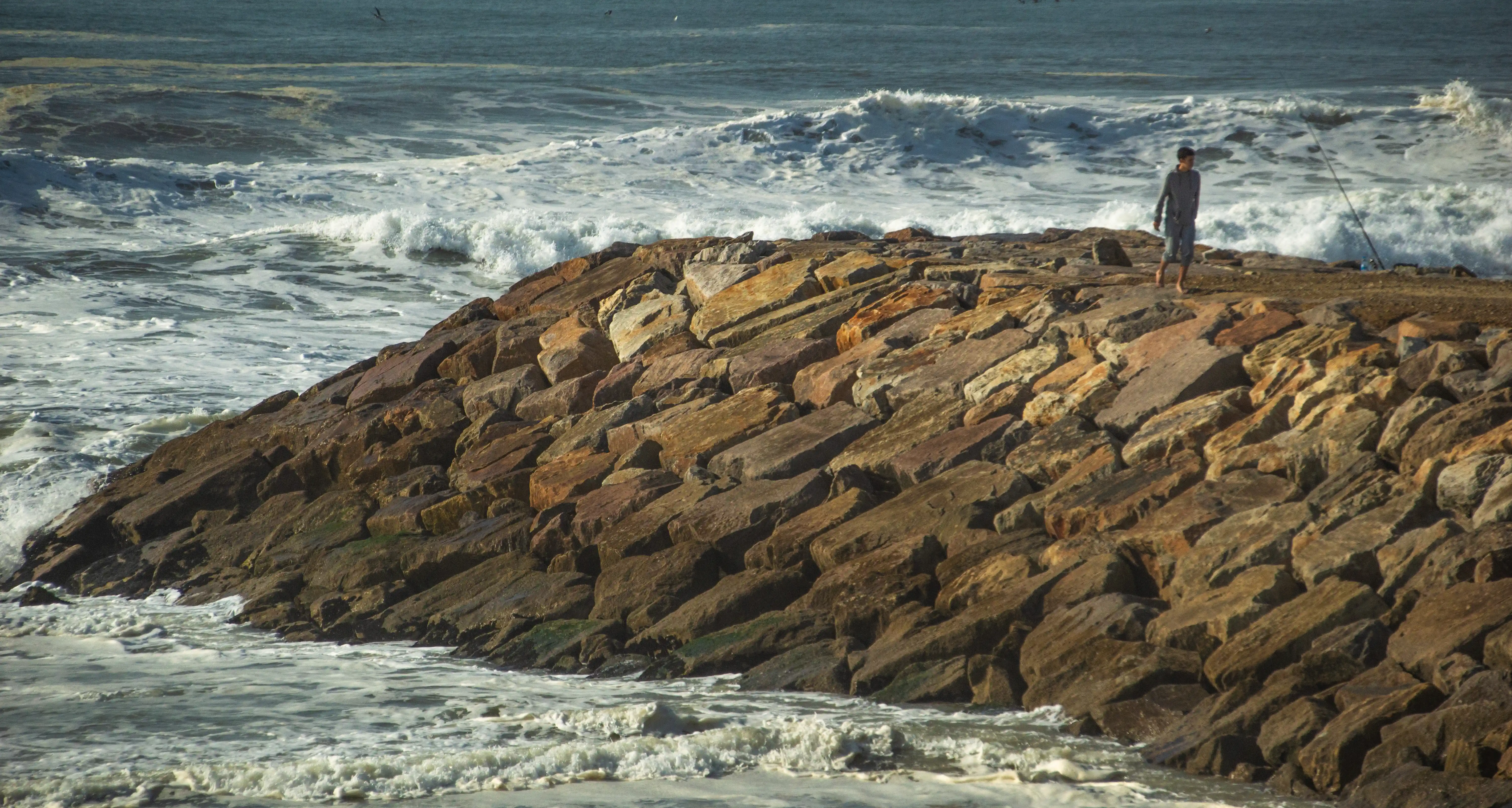A person stands on a stone breakwater, fishing in the ocean as waves crash.