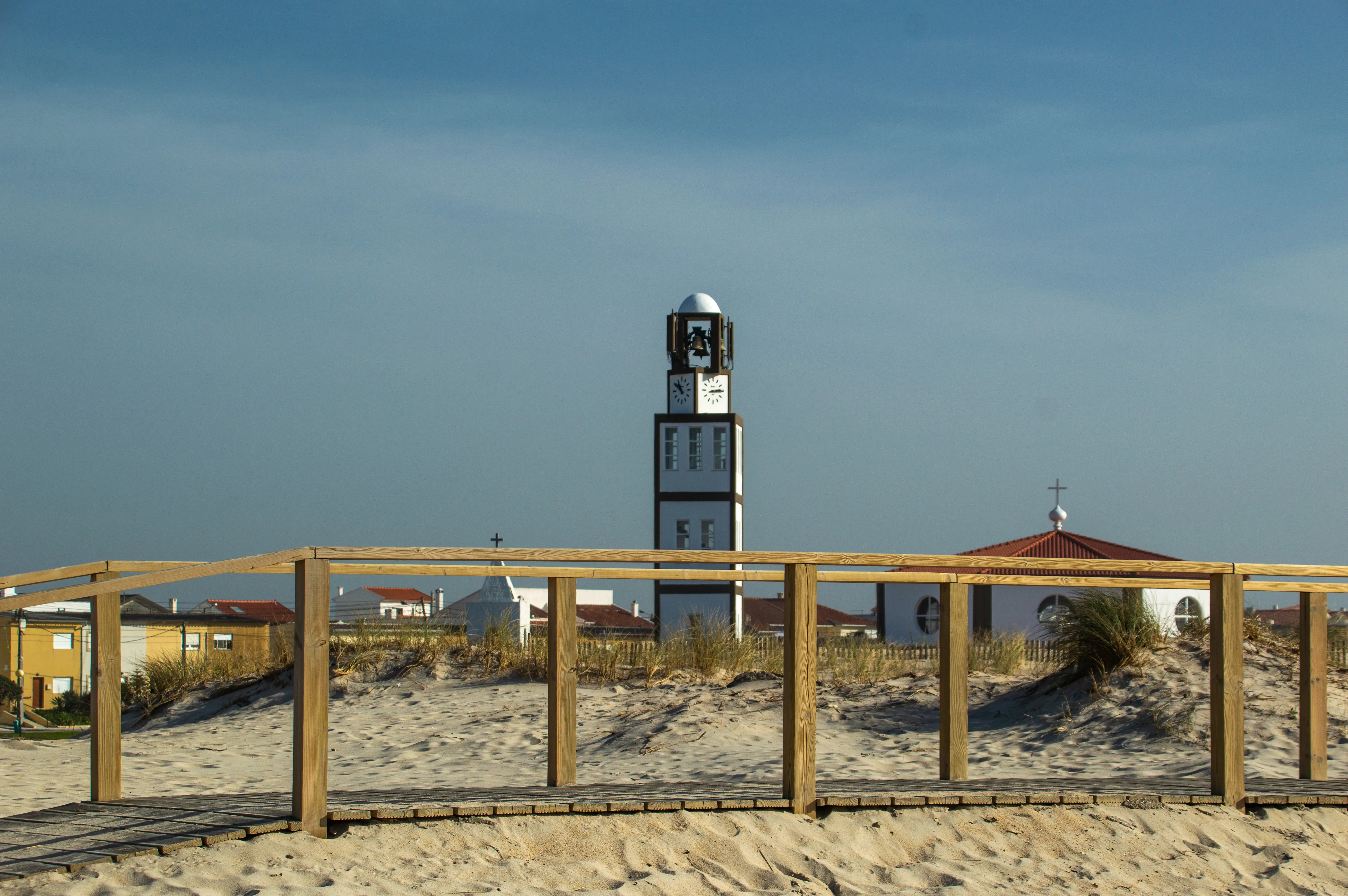 A view across sand dunes and a wooden boardwalk to a modern white and brown bell tower with clocks and a small white chapel with a red roof and a cross, under a clear blue sky.