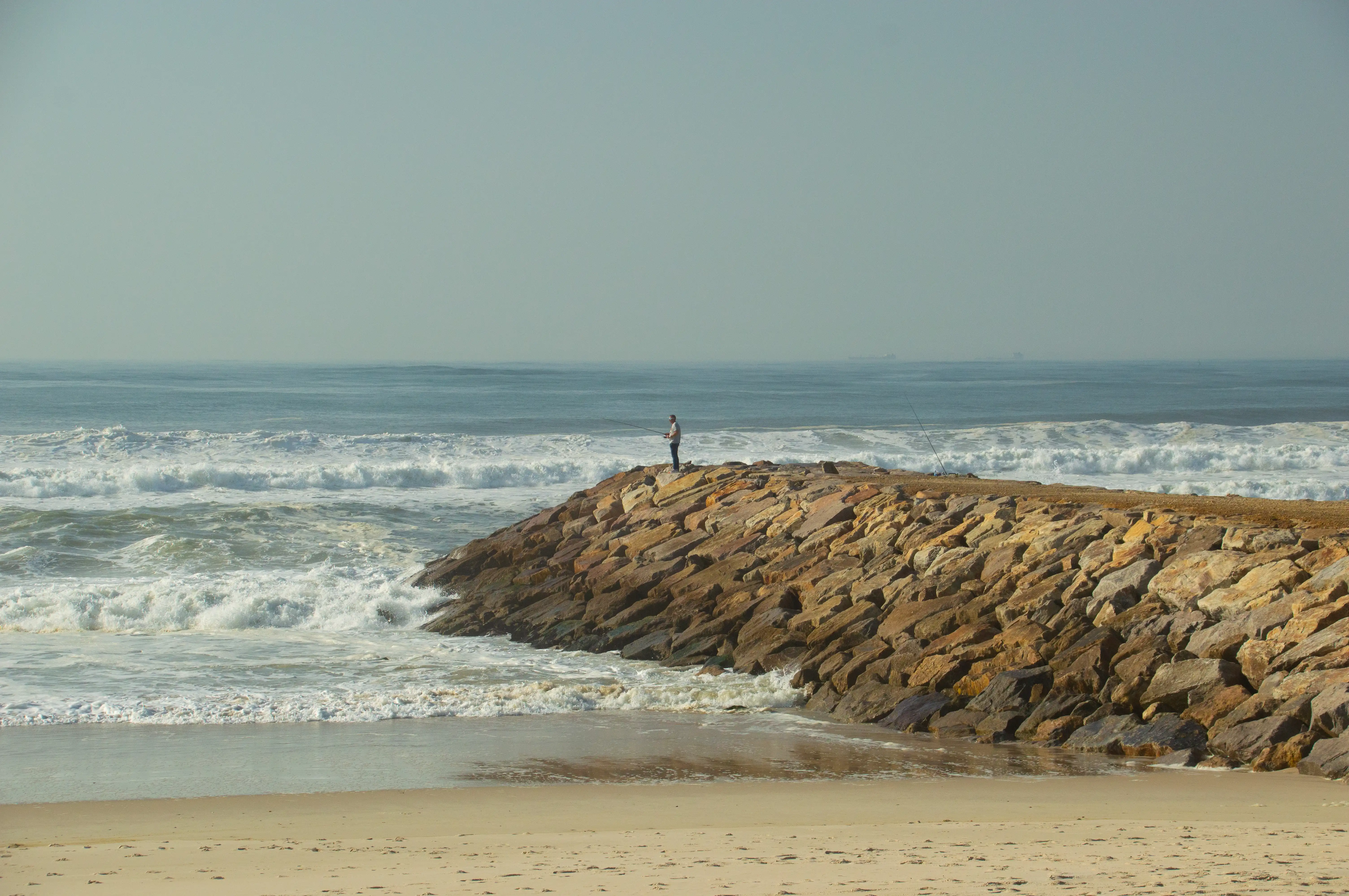 A person fishing from a breakwater made of large, irregular stones, with waves breaking on the shore and the sea stretching to the horizon under a pale sky.