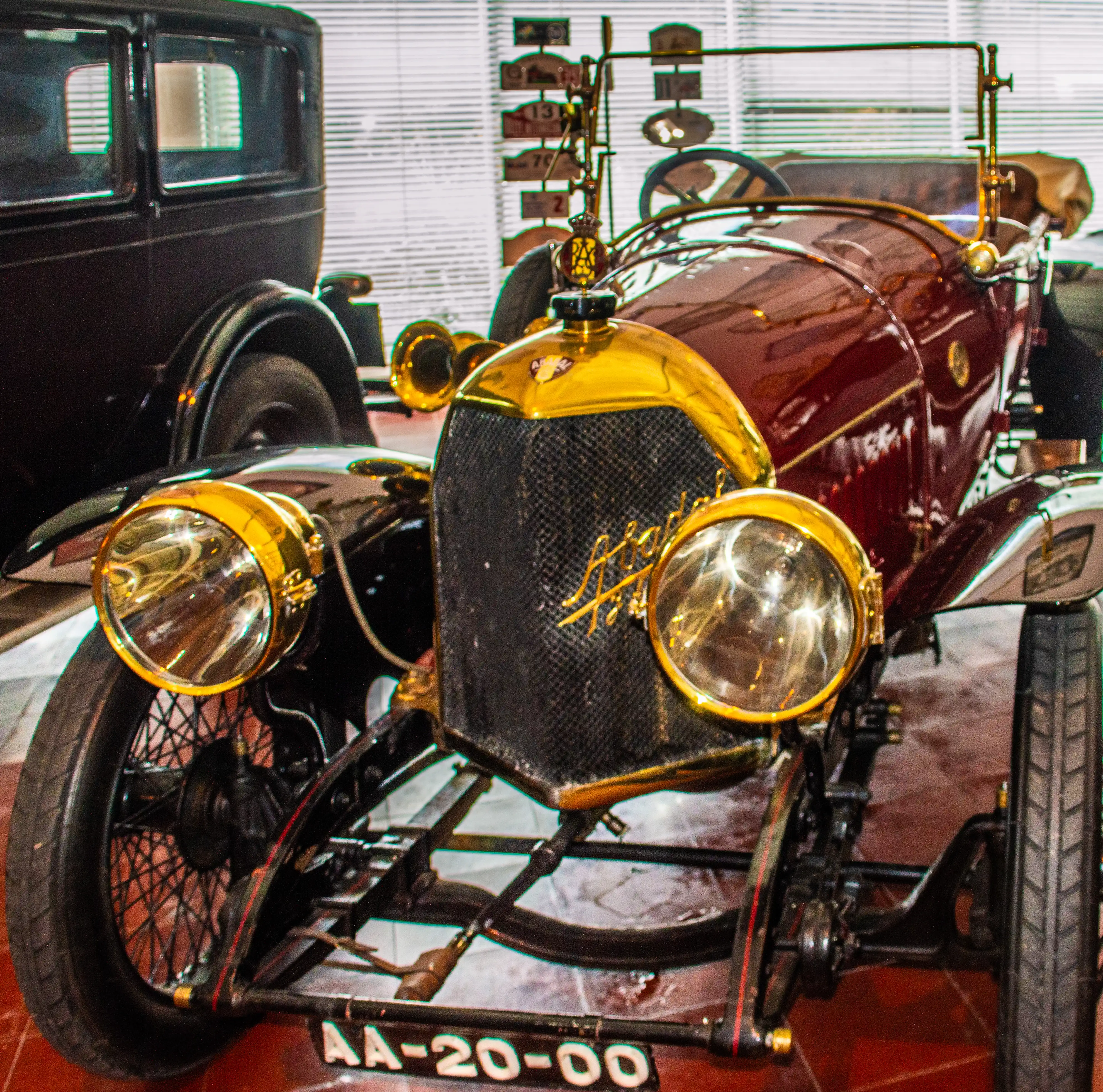 A close-up photograph of a vintage maroon car with a highly polished gold bonnet, grille, and large, circular headlamps. The car has spoked wheels and a license plate that reads AA-20-00.