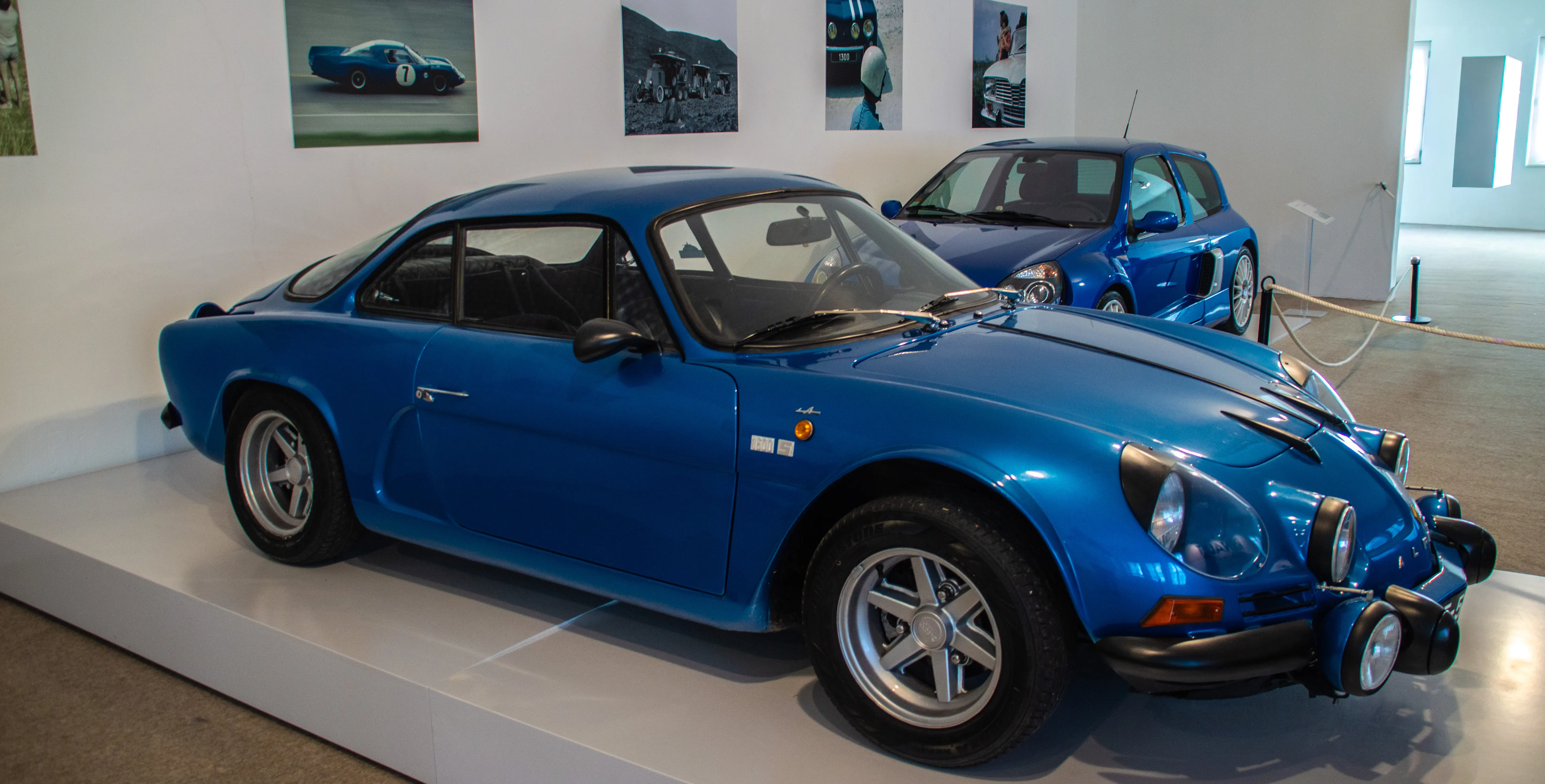 Two blue Alpine sports cars, one classic and one modern, are displayed in a museum with racing photographs on the wall.