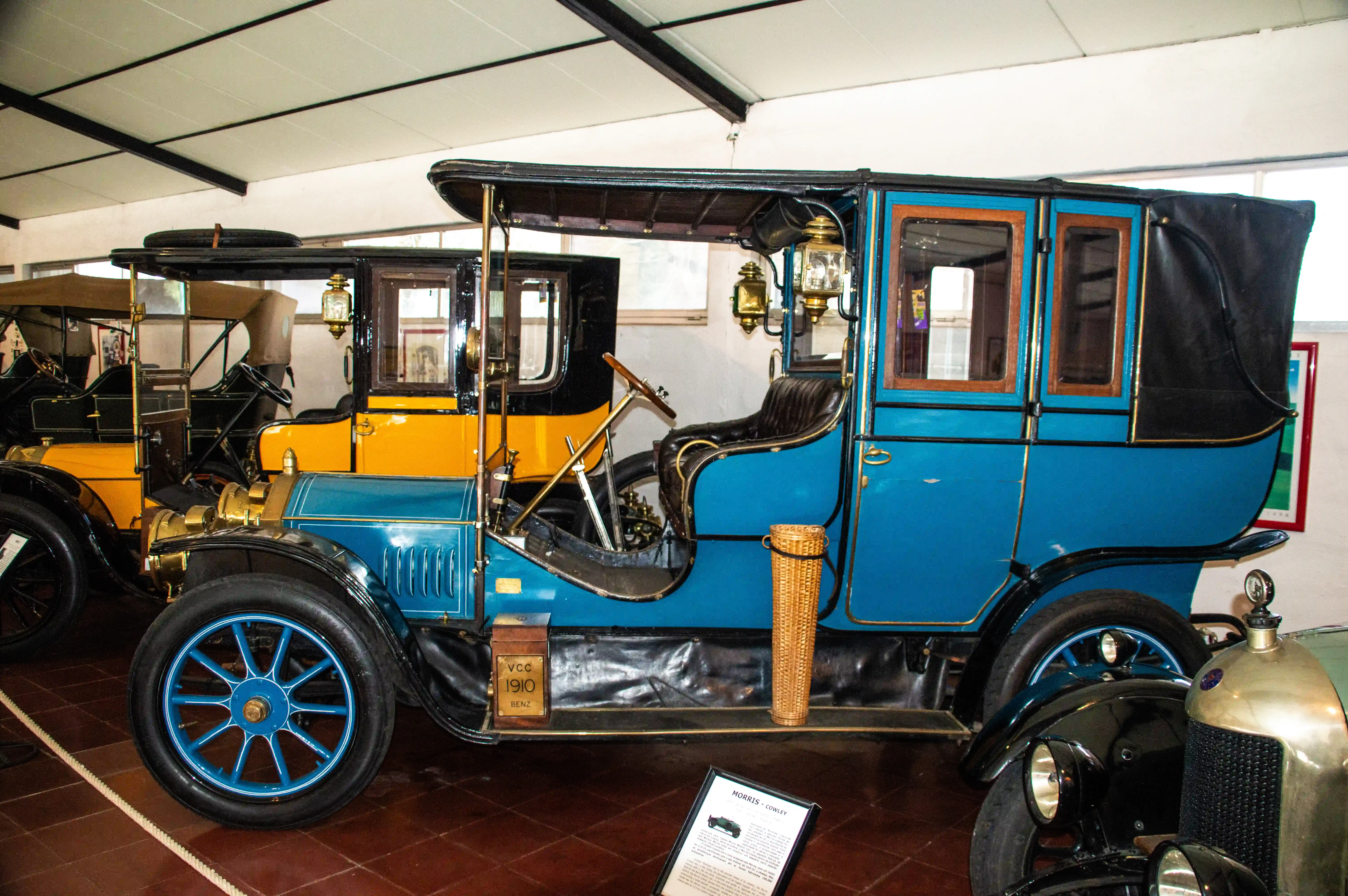 : A striking blue and yellow Benz automobile from 1910 is showcased indoors, possibly in a museum. The vehicle features ornate brass lamps, a wooden-rimmed steering wheel, and a black canvas roof. Another vintage car, primarily yellow and black, is visible behind it, and a portion of a modern-looking green vehicle is in the foreground. A descriptive plaque is placed in front of the Benz.