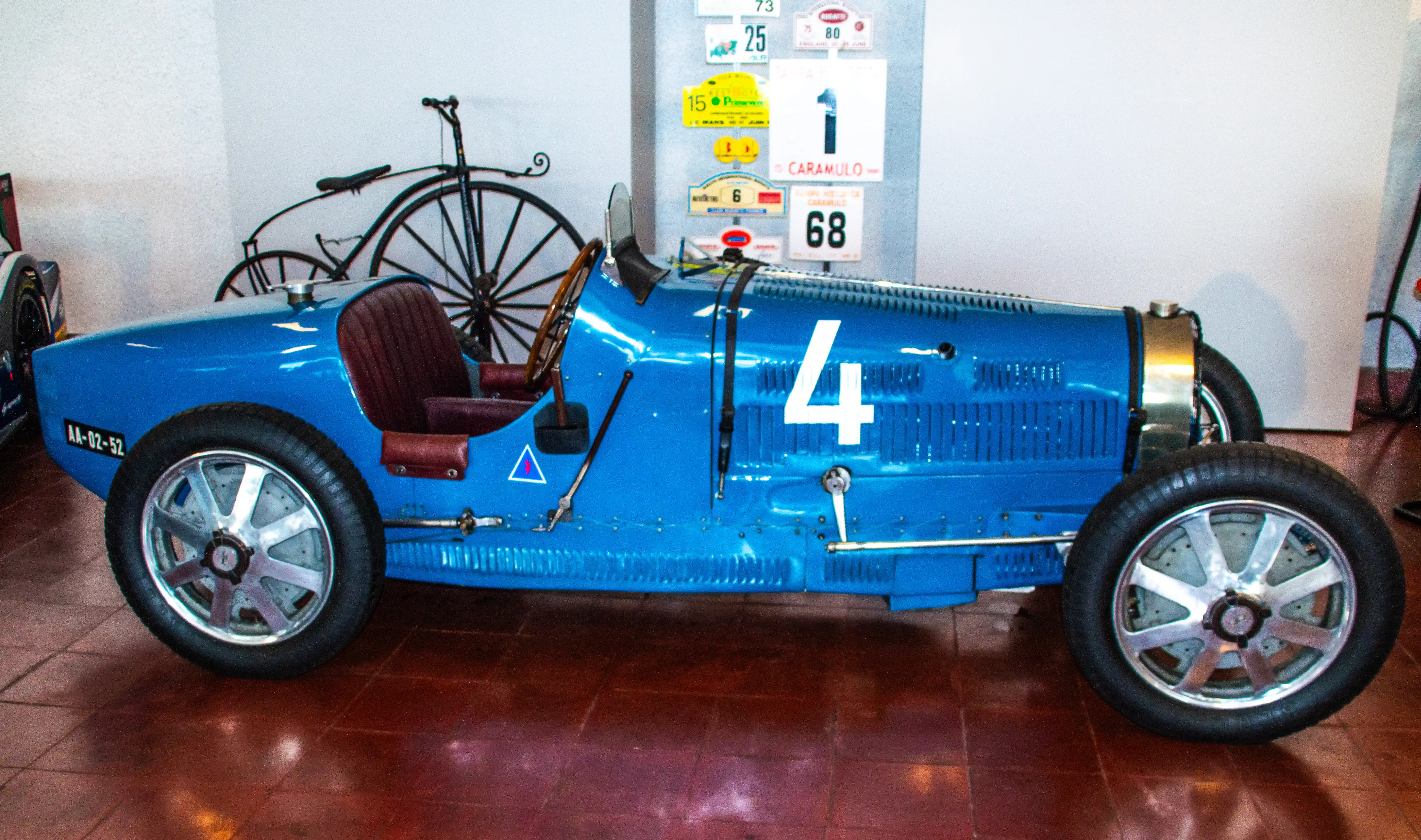 Side view of a bright blue vintage Bugatti racing car with the number 4 prominently displayed on its bodywork. The car has a classic open cockpit, leather seats, and silver spoked wheels. A penny-farthing bicycle and rally plaques are visible in the background.
