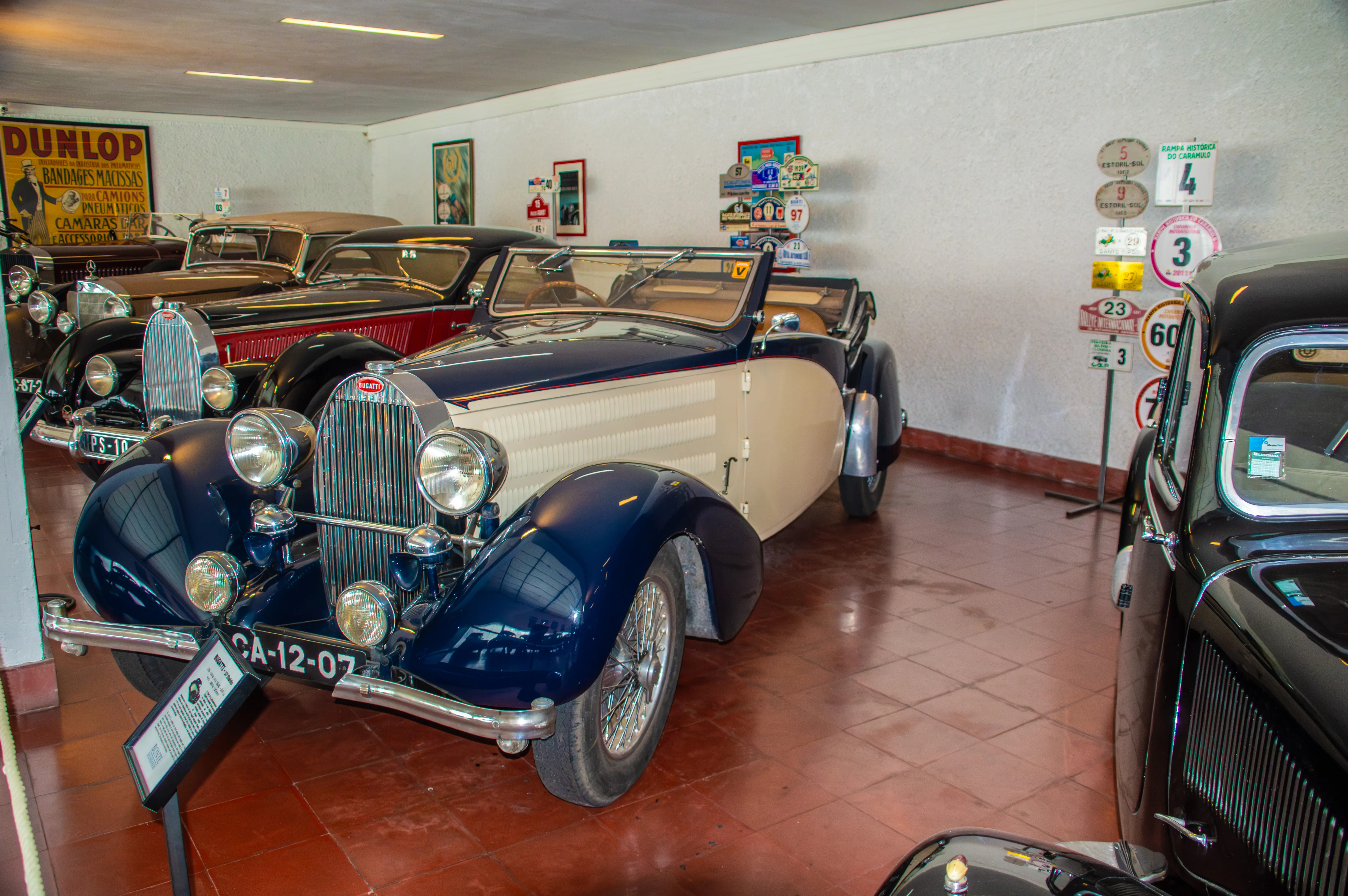 A vintage dark blue and cream Bugatti convertible with wire-spoke wheels, parked in a showroom with other classic cars and historical racing signs on the wall.