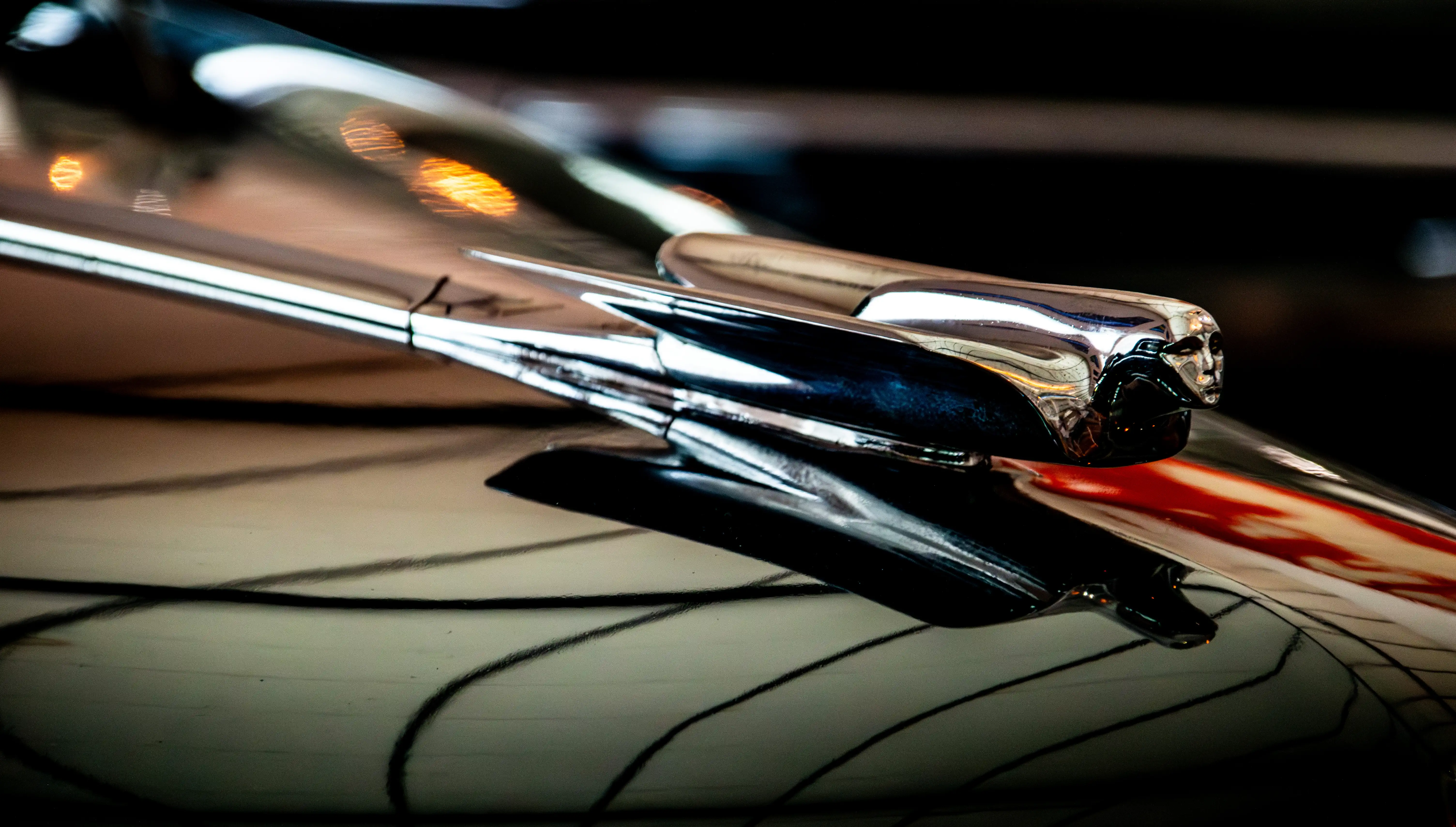 A detailed, close-up photograph of a vintage car's chrome hood ornament shaped like a human face, set against a highly reflective car bonnet.