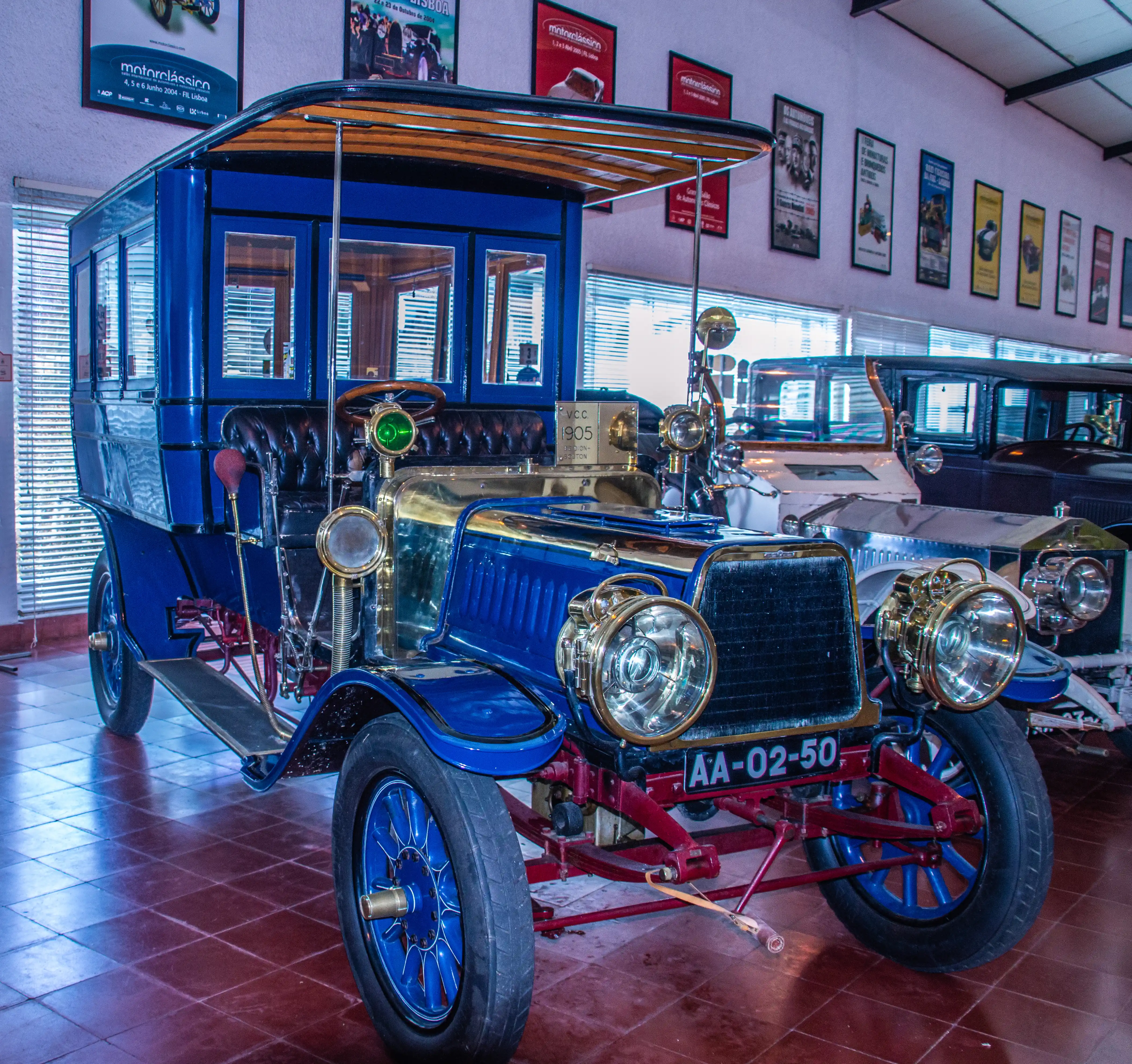 A bright blue 1905 motorcar with brass trim, large circular headlamps, and a wooden canopy roof, parked on a tiled floor with other vintage cars in the background.