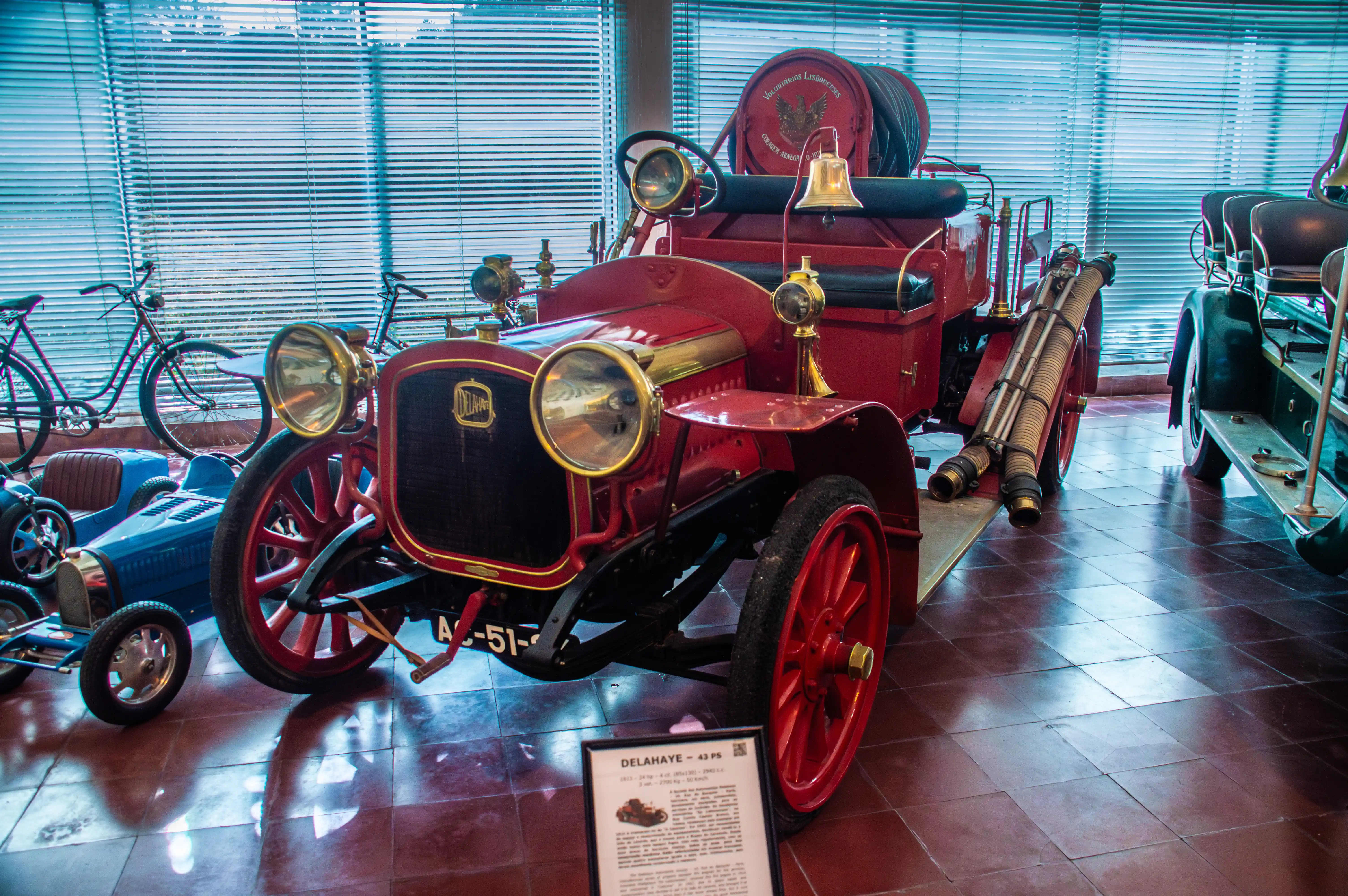 A close-up shot of a red vintage Delahaye fire engine with brass accents, a bell, and coiled hoses. The vehicle is on display in a museum, with other antique vehicles visible in the background.