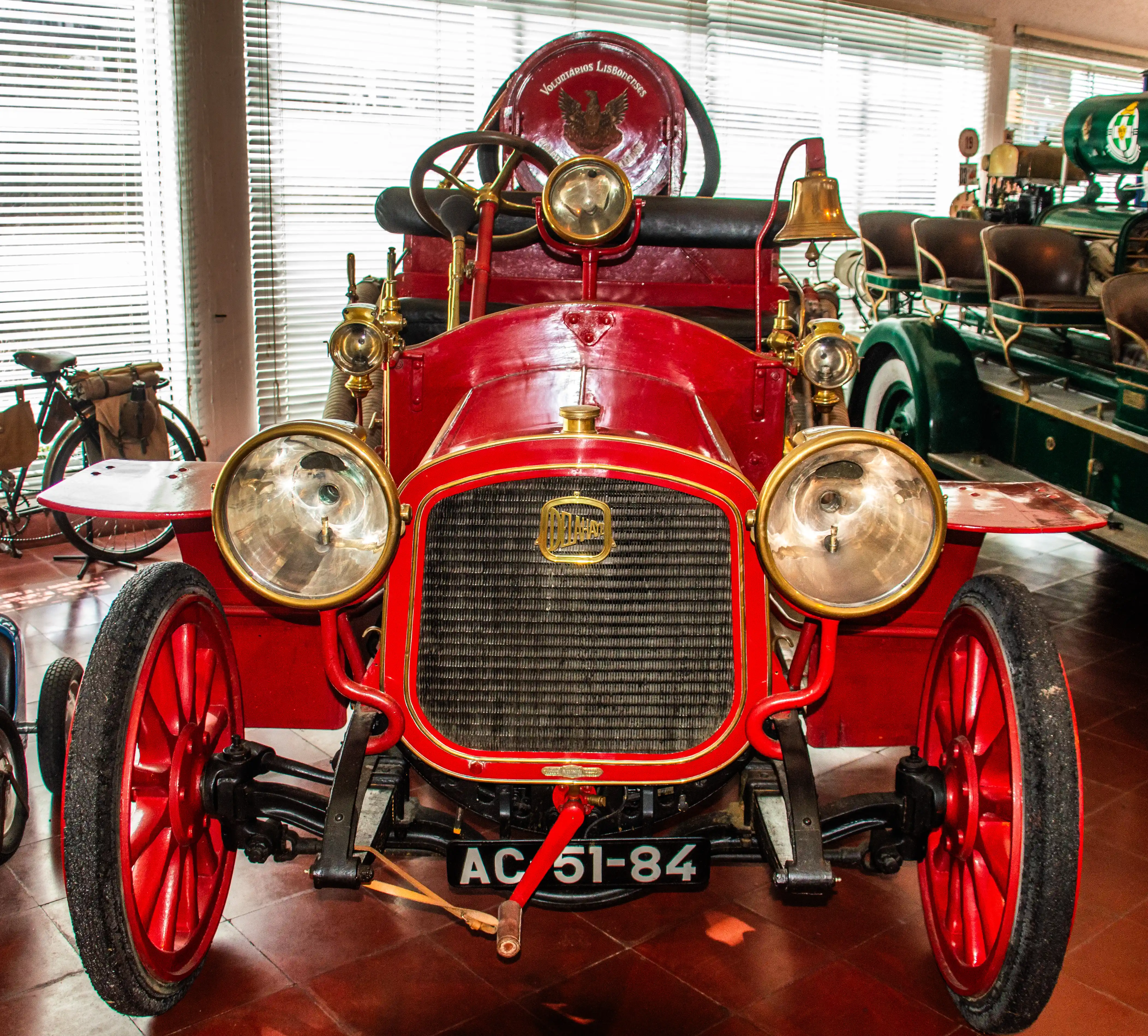 Front view of a red vintage fire engine with brass detailing, a bell, and the license plate AC 51-84, displayed indoors with a green fire engine and a bicycle in the background.