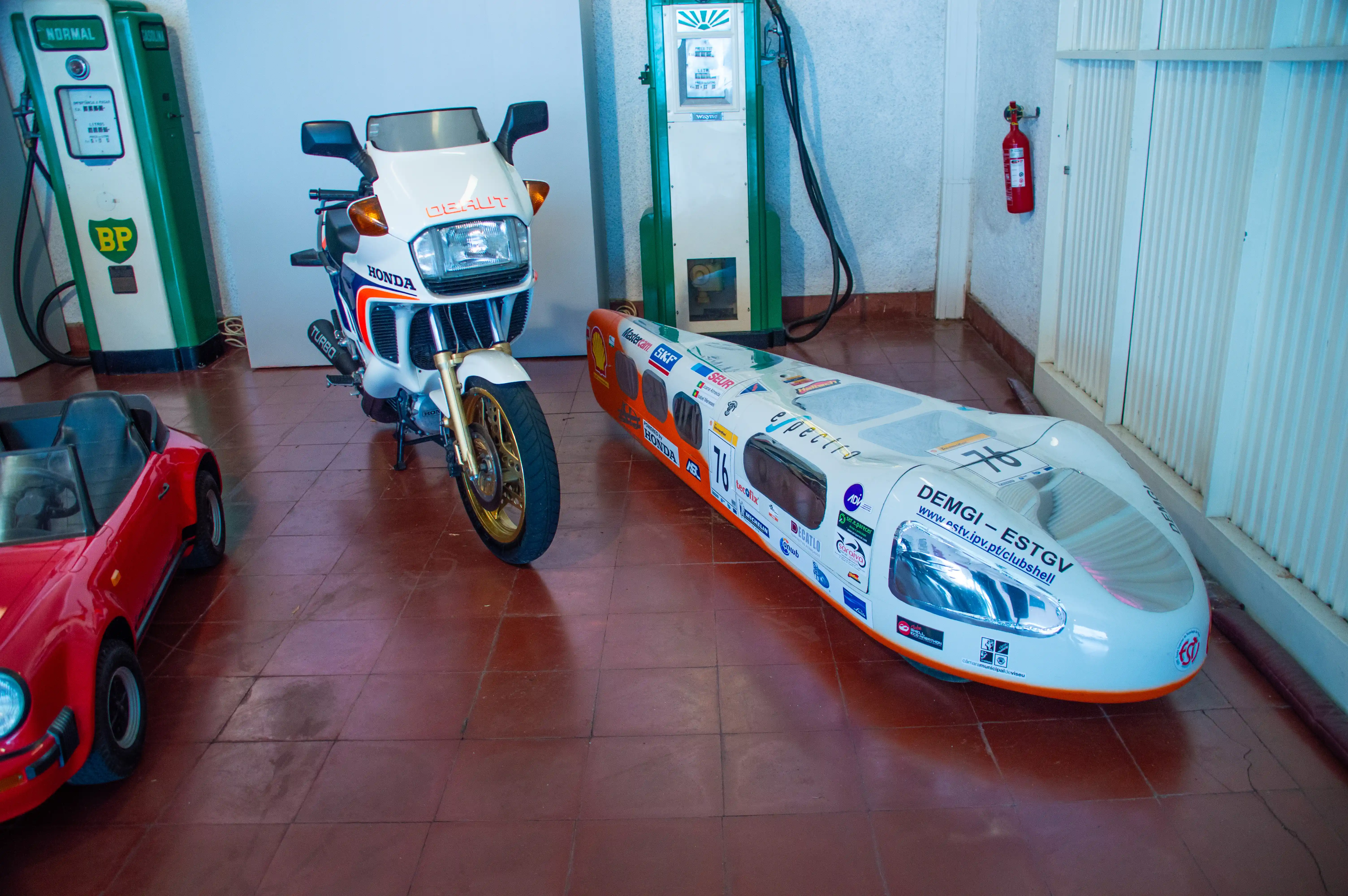 A red car, a white Honda motorcycle and a white, orange, and silver aerodynamic streamliner vehicle are parked in a garage next to vintage petrol pumps.