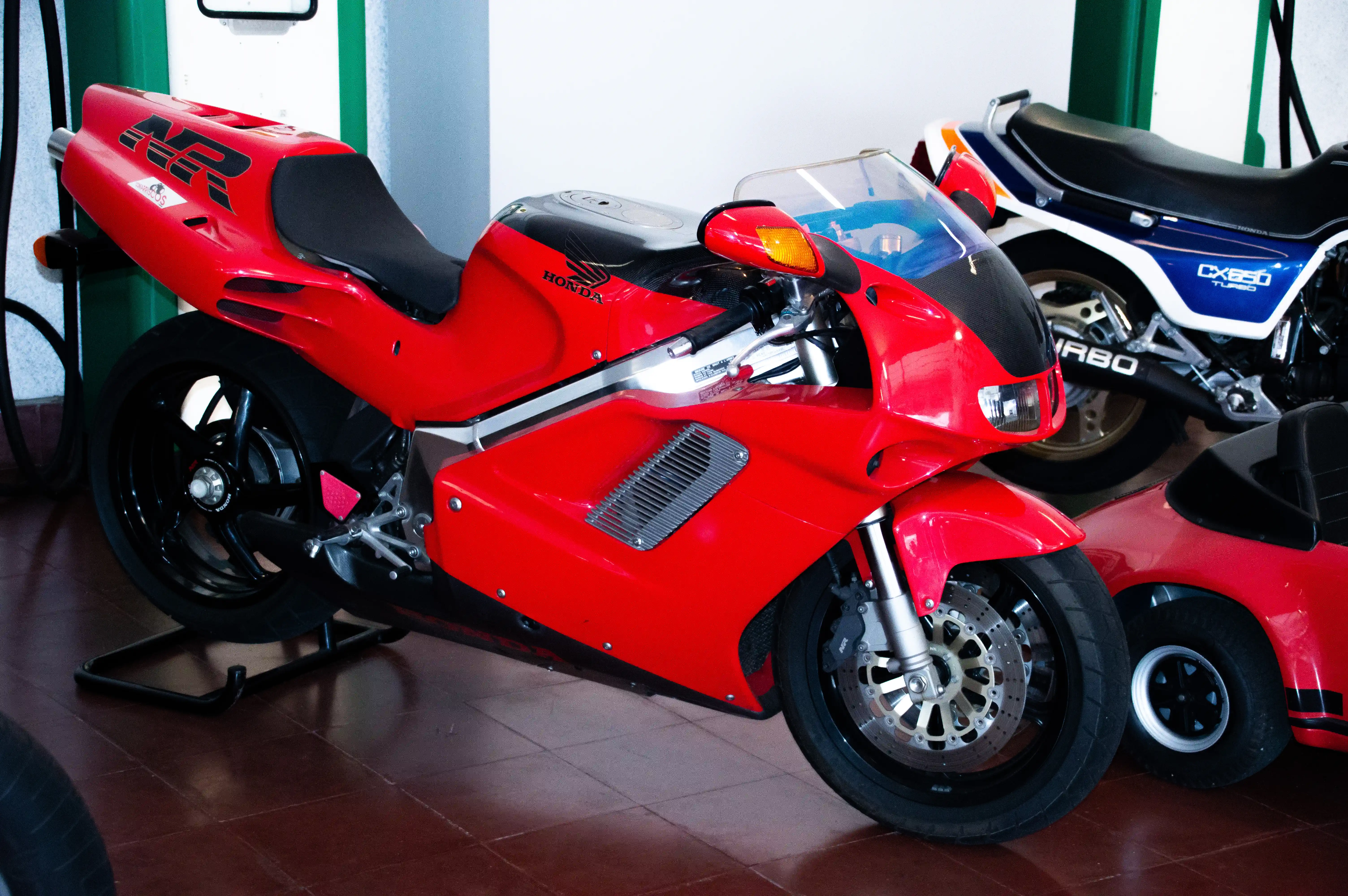 A bright red Honda NR motorcycle with black accents and a clear windshield is parked on a dark tiled floor. In the background, a blue and white Honda CX650 Turbo motorcycle is visible.