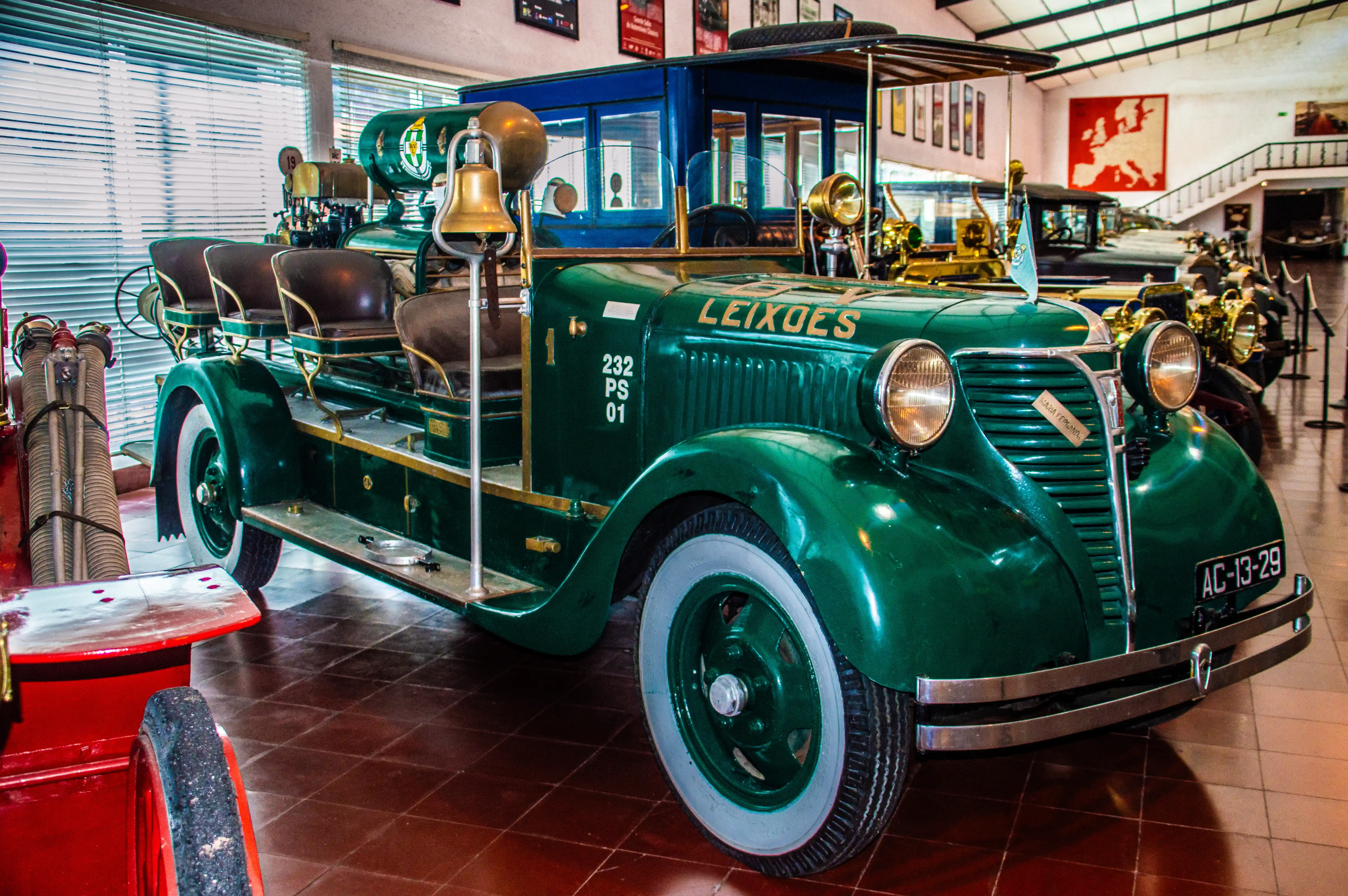 A shiny, emerald green vintage fire engine with gold trim and seating for multiple passengers, parked indoors with other classic vehicles in the background