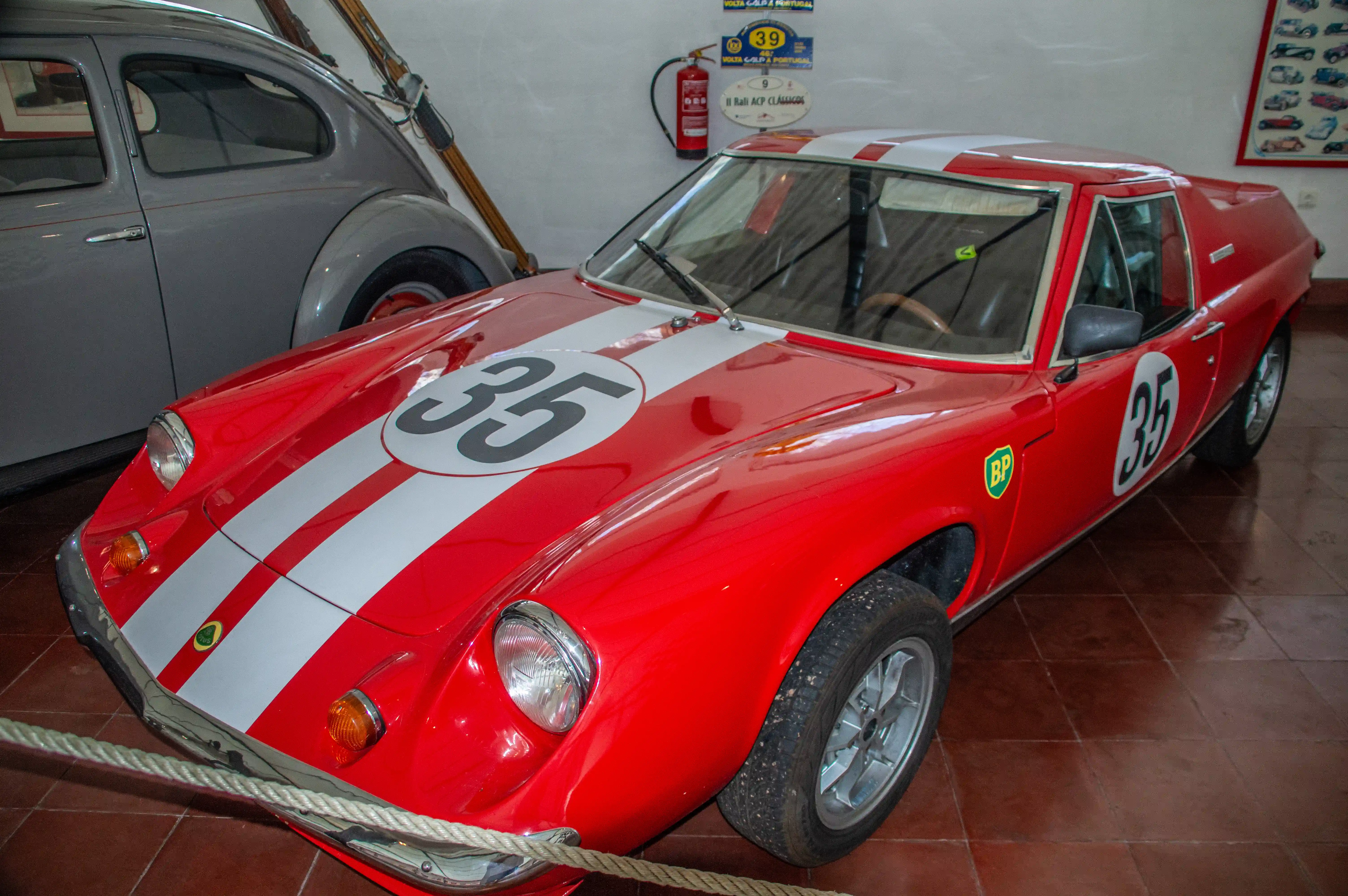A red Lotus Europa racing car with white stripes and the number 35 on the bonnet and door, parked indoors next to a grey car.