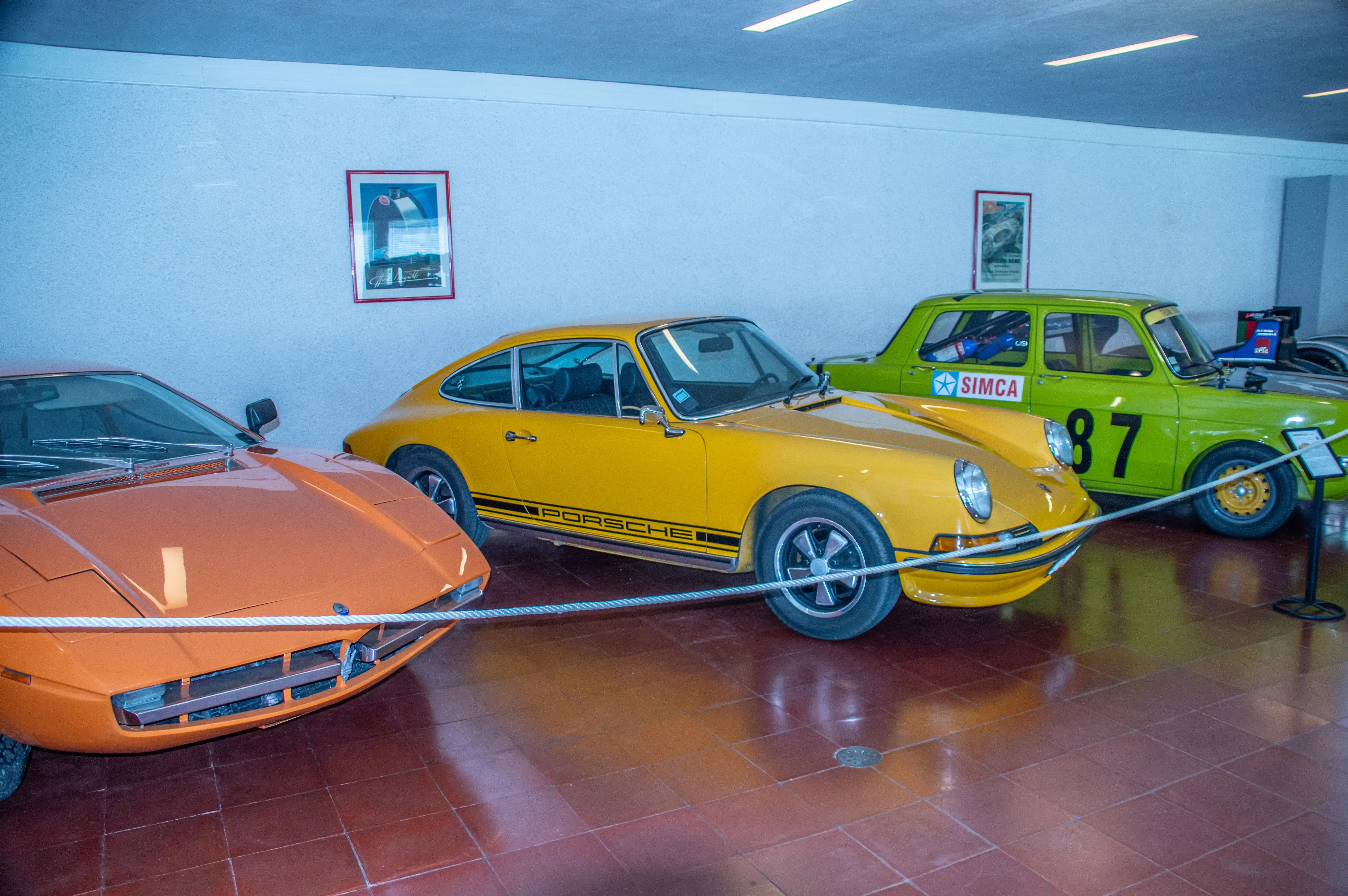 A collection of vintage cars including an orange Maserati, a yellow Porsche, and a green Simca race car with the number 87 are displayed indoors, with framed pictures on the wall and a tiled floor.
