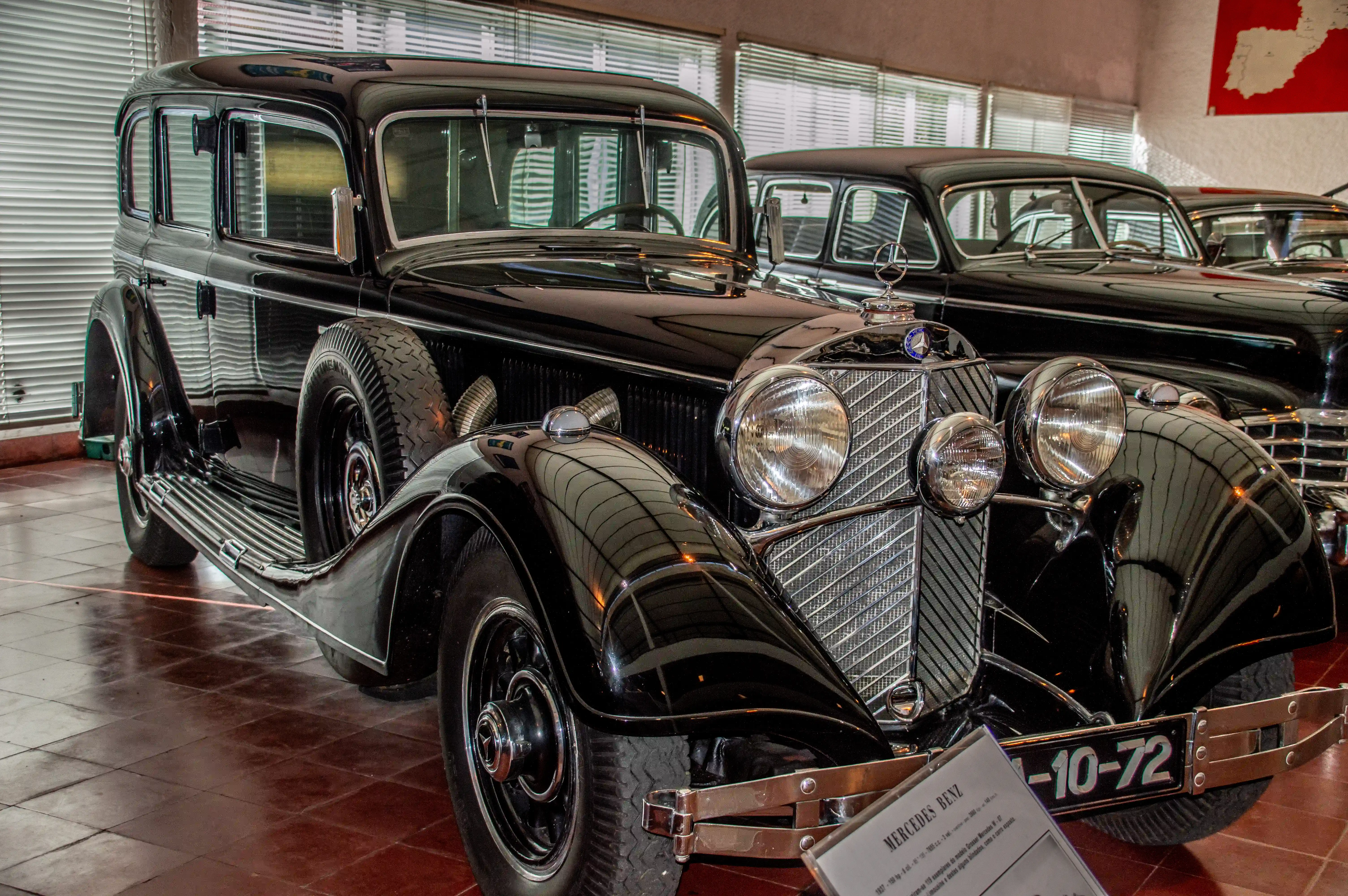 A polished black vintage Mercedes-Benz sedan, with a prominent chrome grille and round headlights, is parked indoors. The car has a spare tyre mounted on its left side.