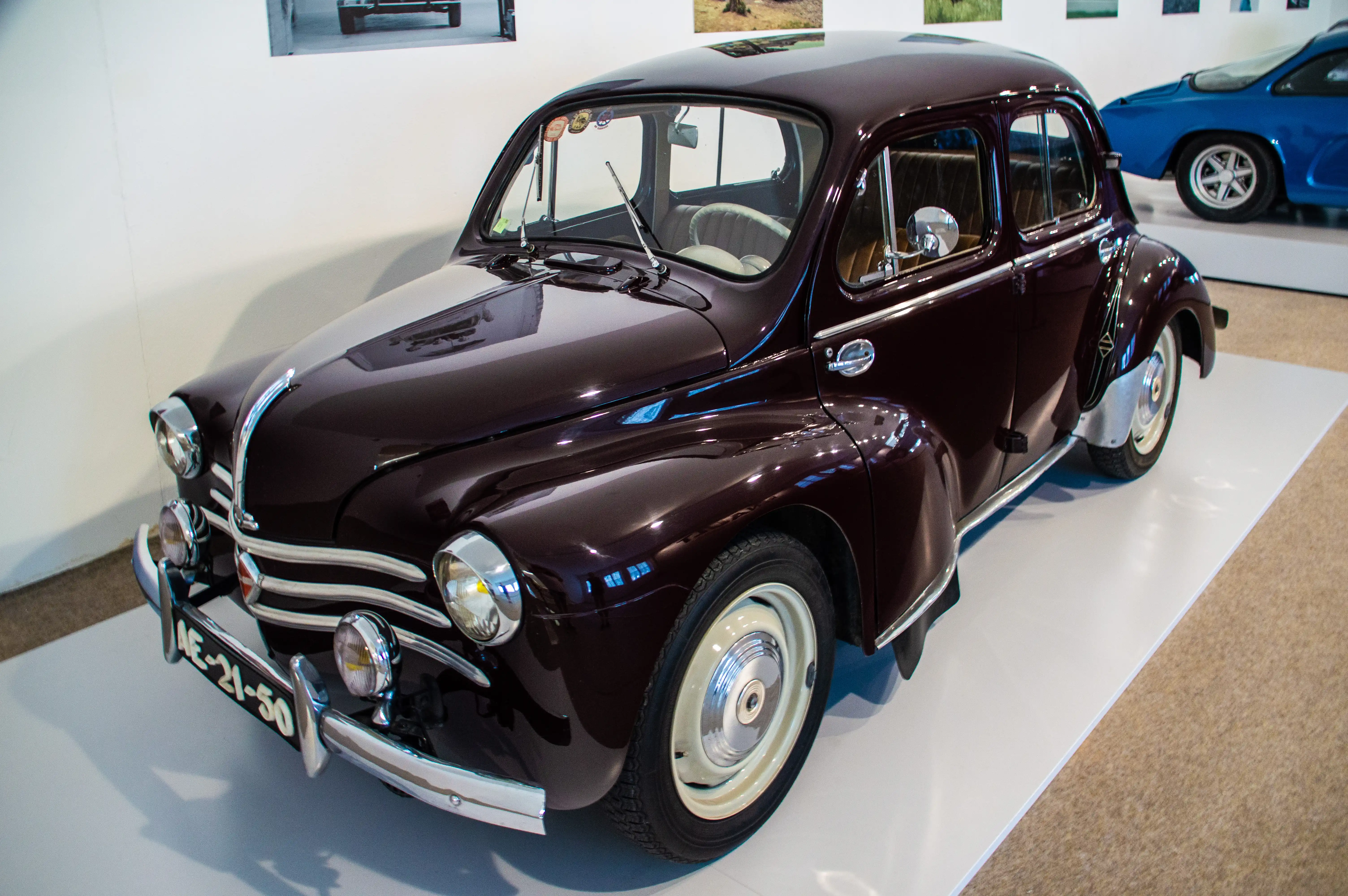 A vintage deep burgundy Renault Dauphine car from the front-right, parked on a white display surface with a blue sports car in the background.