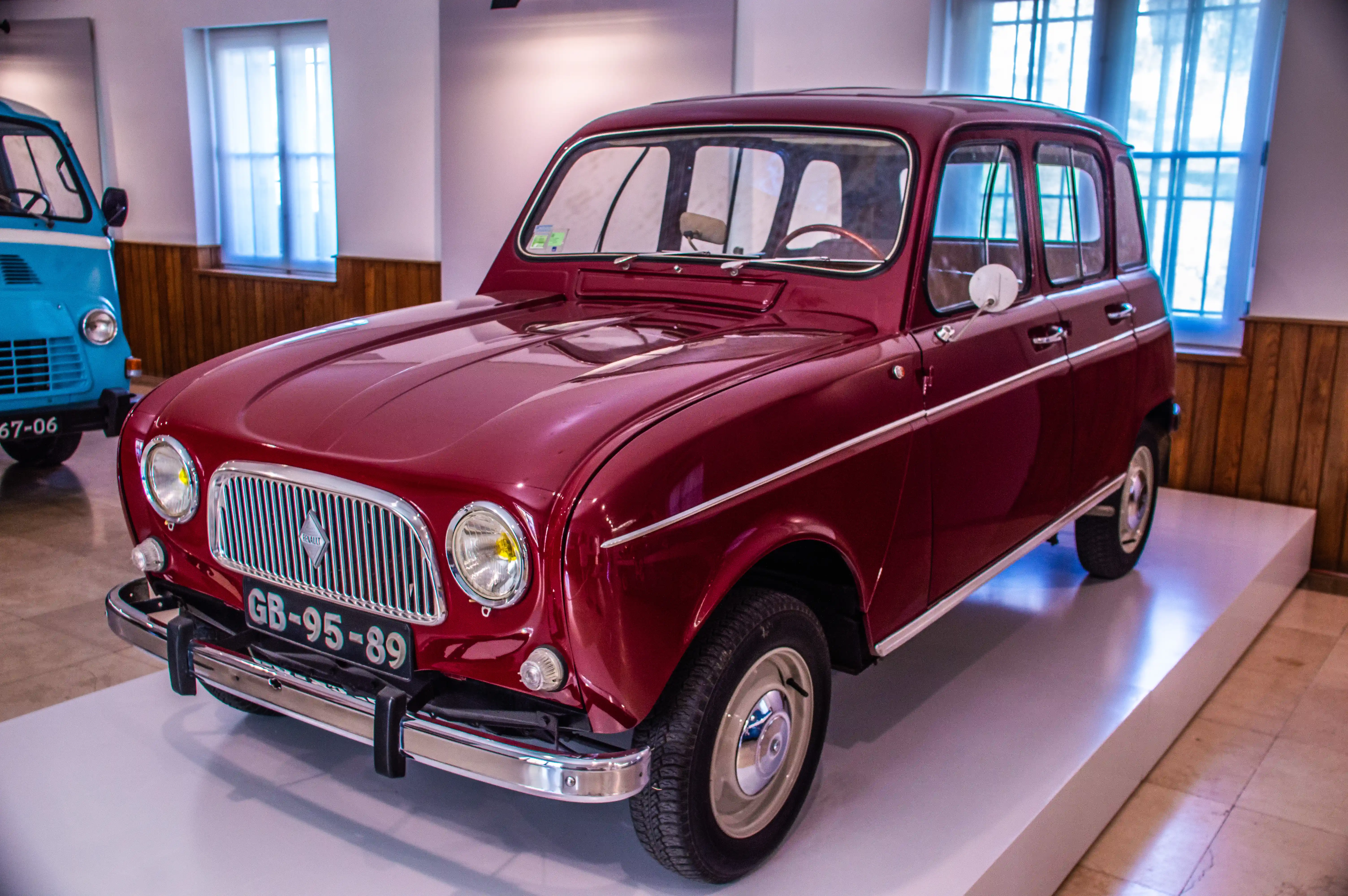 A burgundy Renault 4 car from the 1960s or 1970s is parked on a white platform in a museum. The car has a chrome grille and a license plate reading 