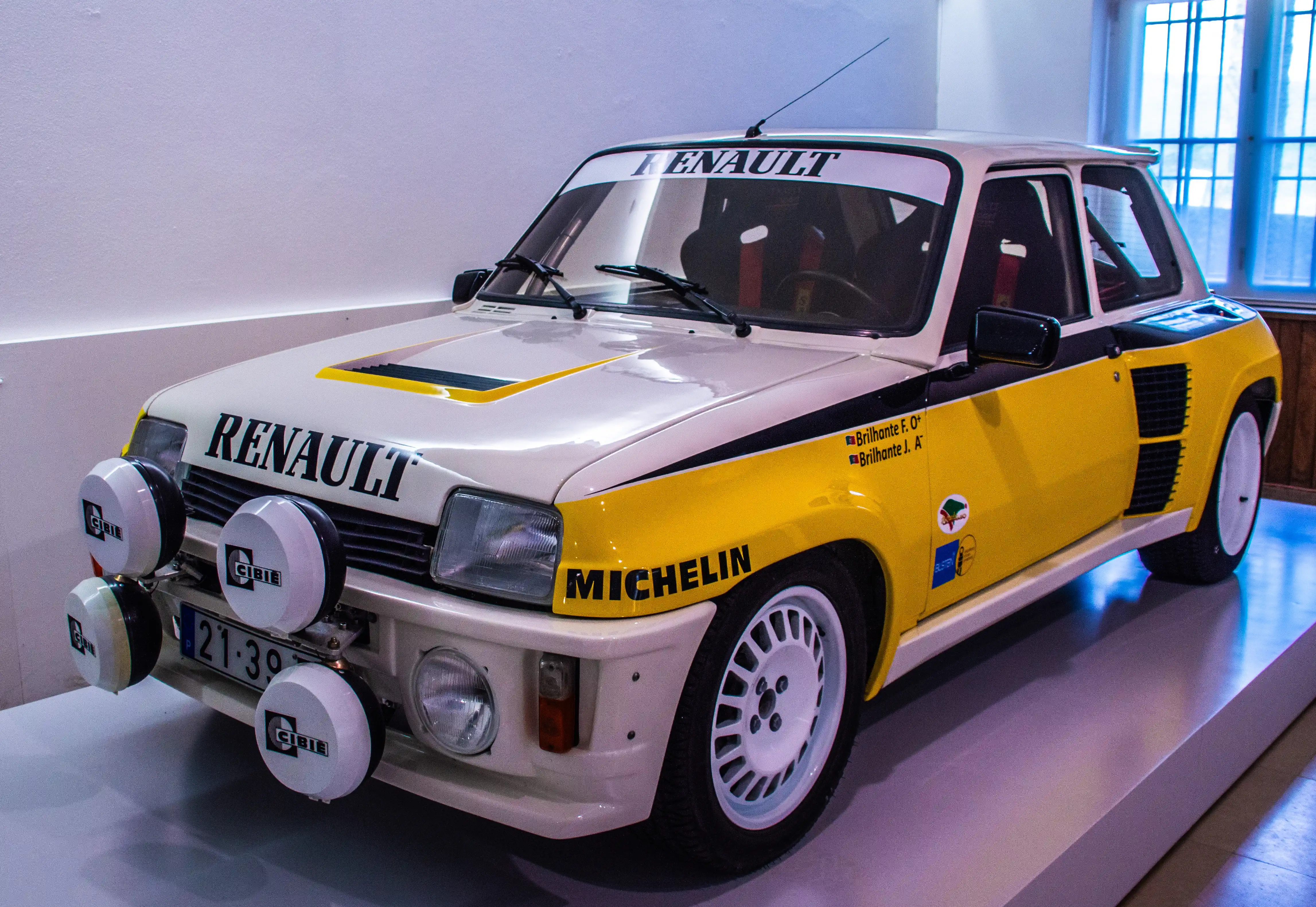 A front-three-quarter view of a classic white and yellow Renault 5 Turbo rally car on display. It has large Cibie fog lights, a 