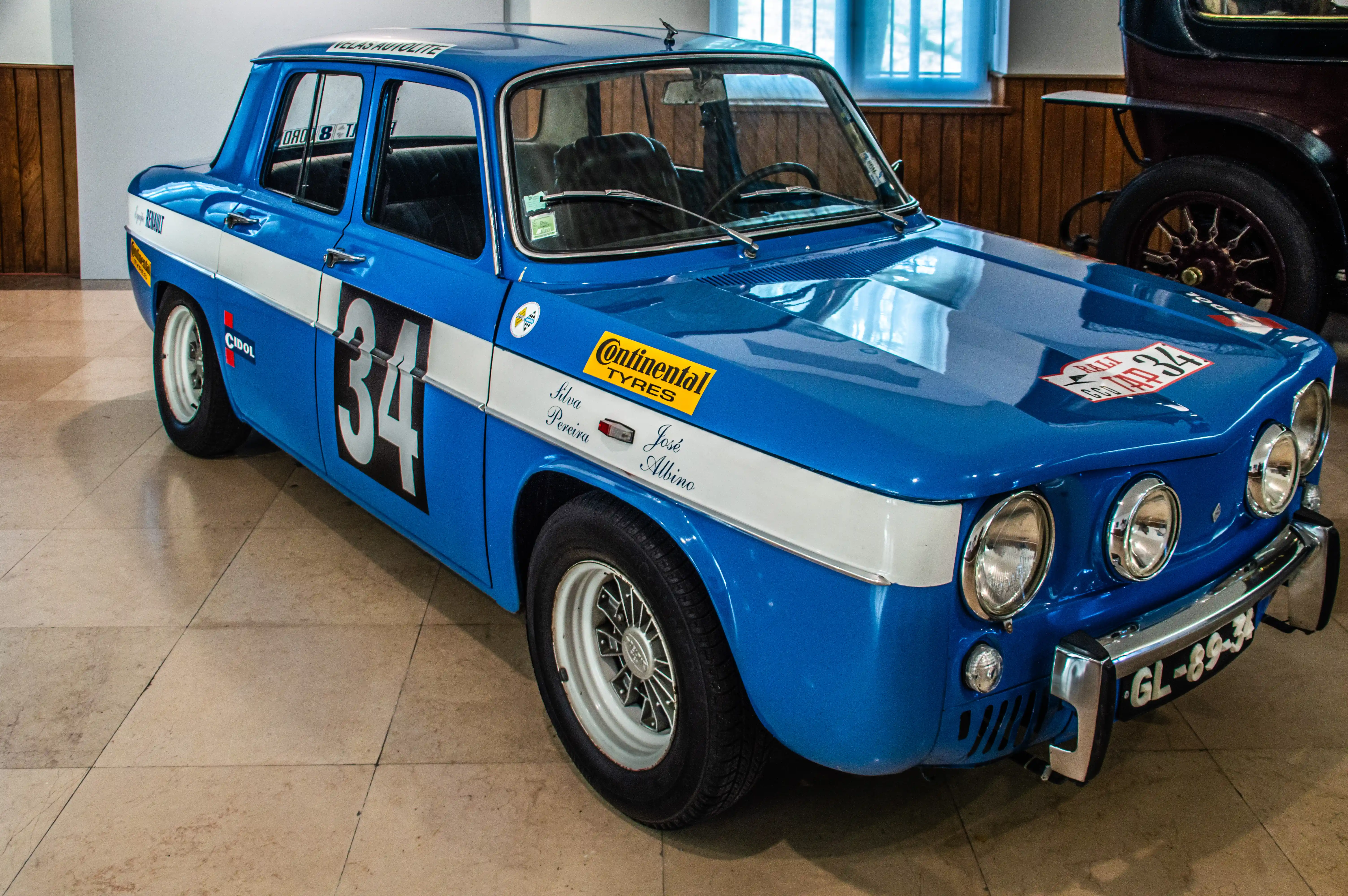 A classic blue rally car with white racing stripes, number 34 on the door, and sponsor logos, showcased indoors.