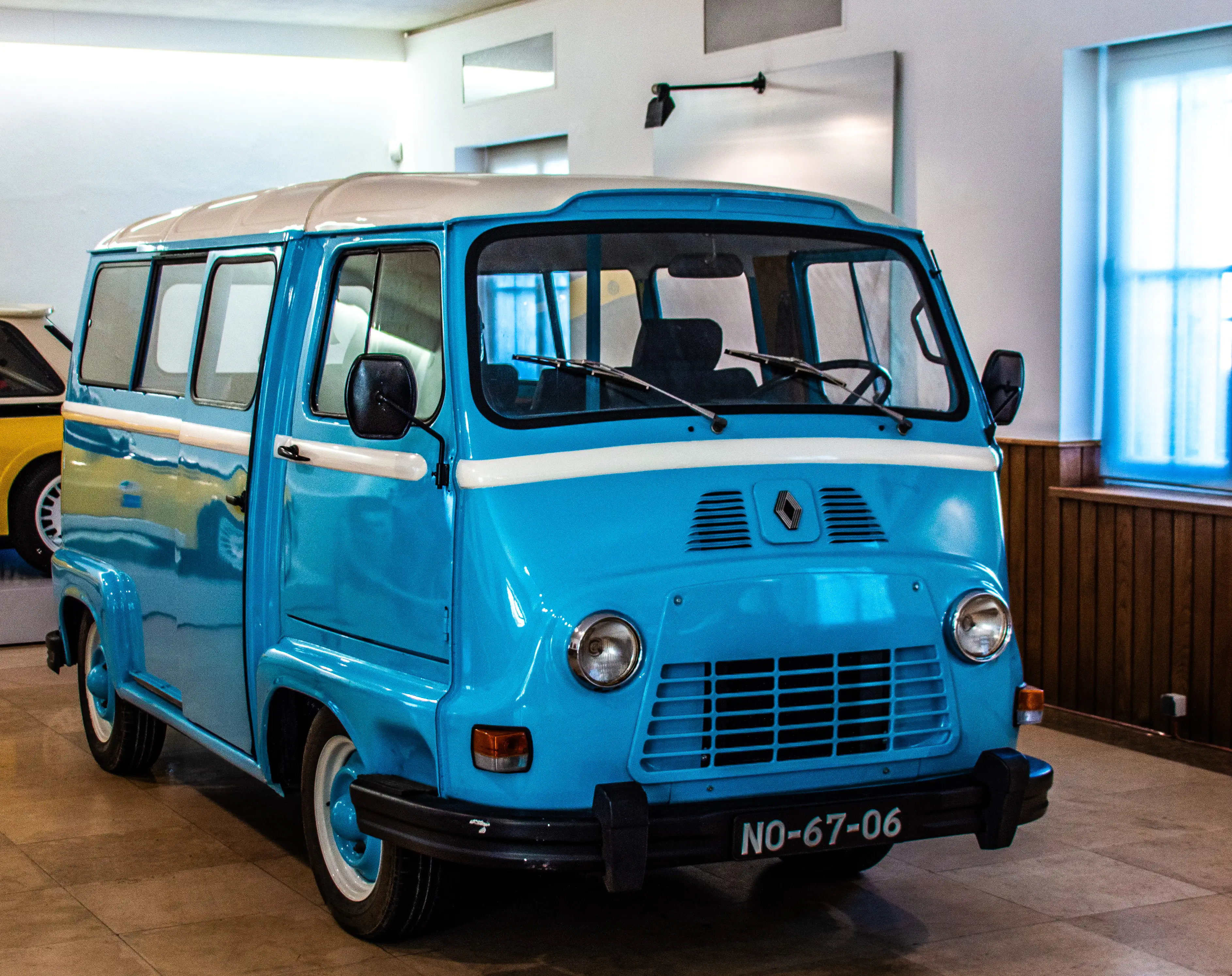 A bright blue and white vintage Renault Estafette van with a yellow stripe, parked indoors. The van has a black bumper with a license plate 