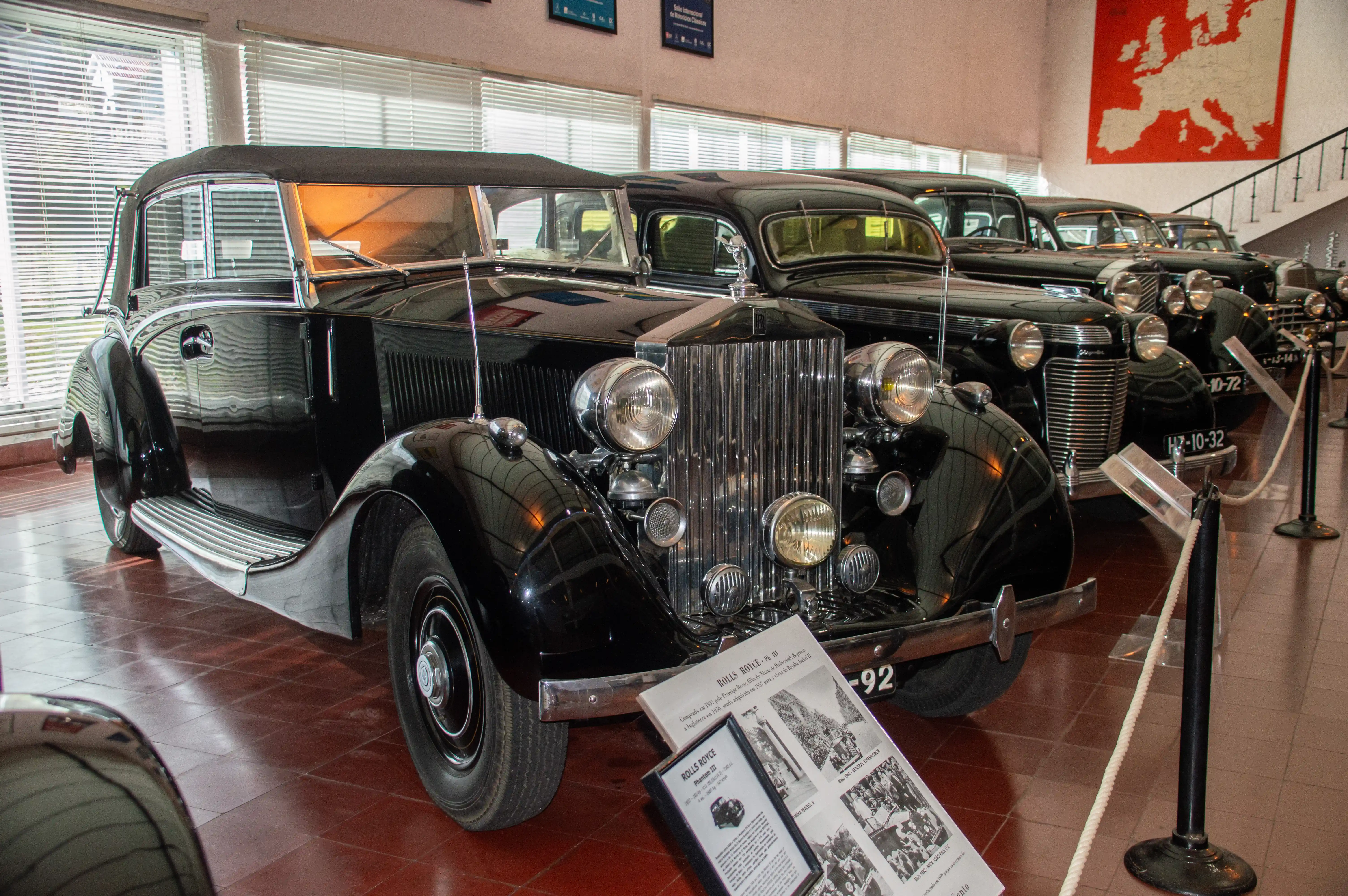  Close-up view of a gleaming black Rolls-Royce Phantom III convertible in a museum, with other vintage Rolls-Royce cars and a map of Europe in the background.