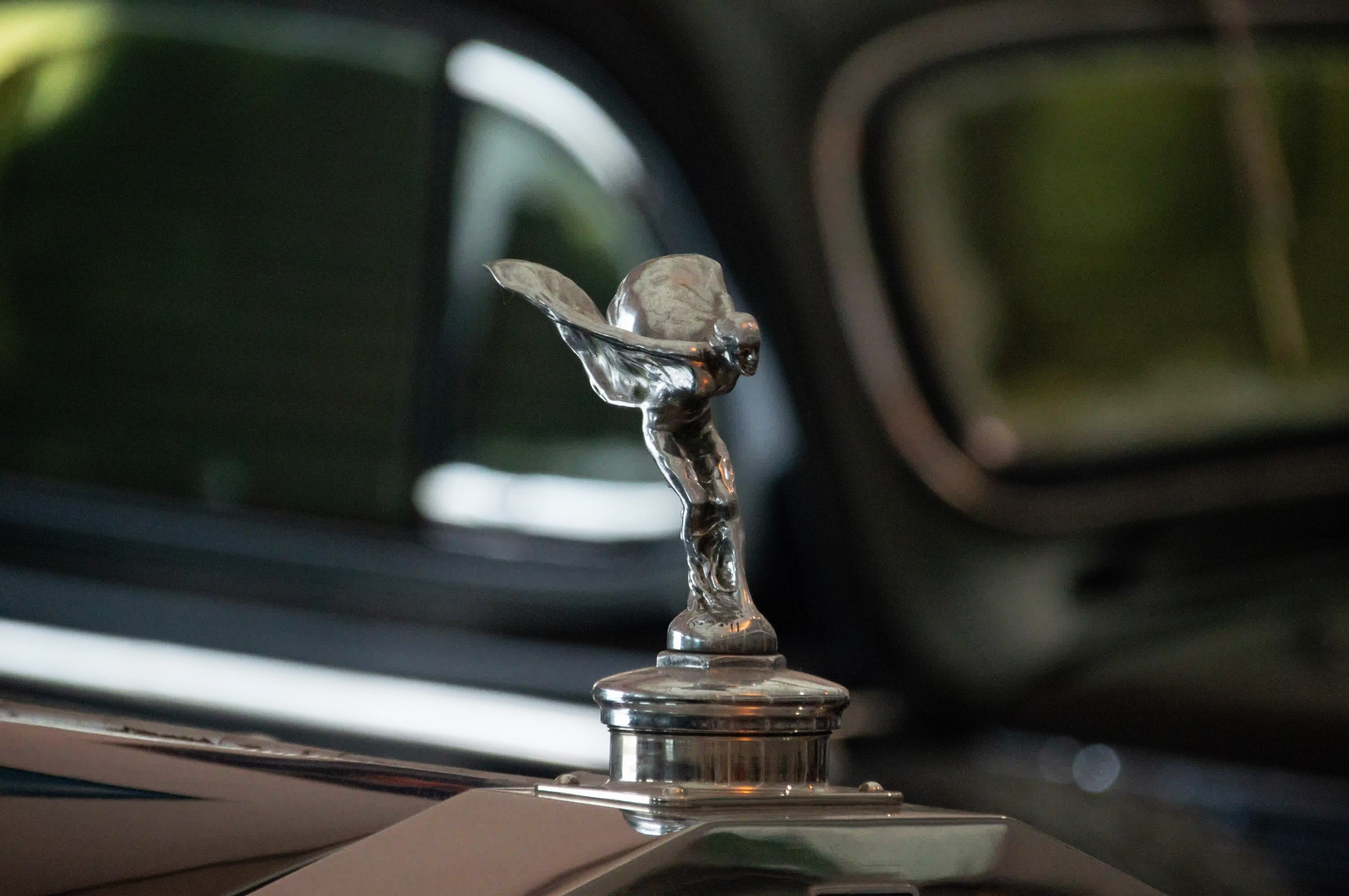 Close-up of the chrome 'Spirit of Ecstasy' mascot on a car bonnet, with a blurred background of the car's interior and windows.