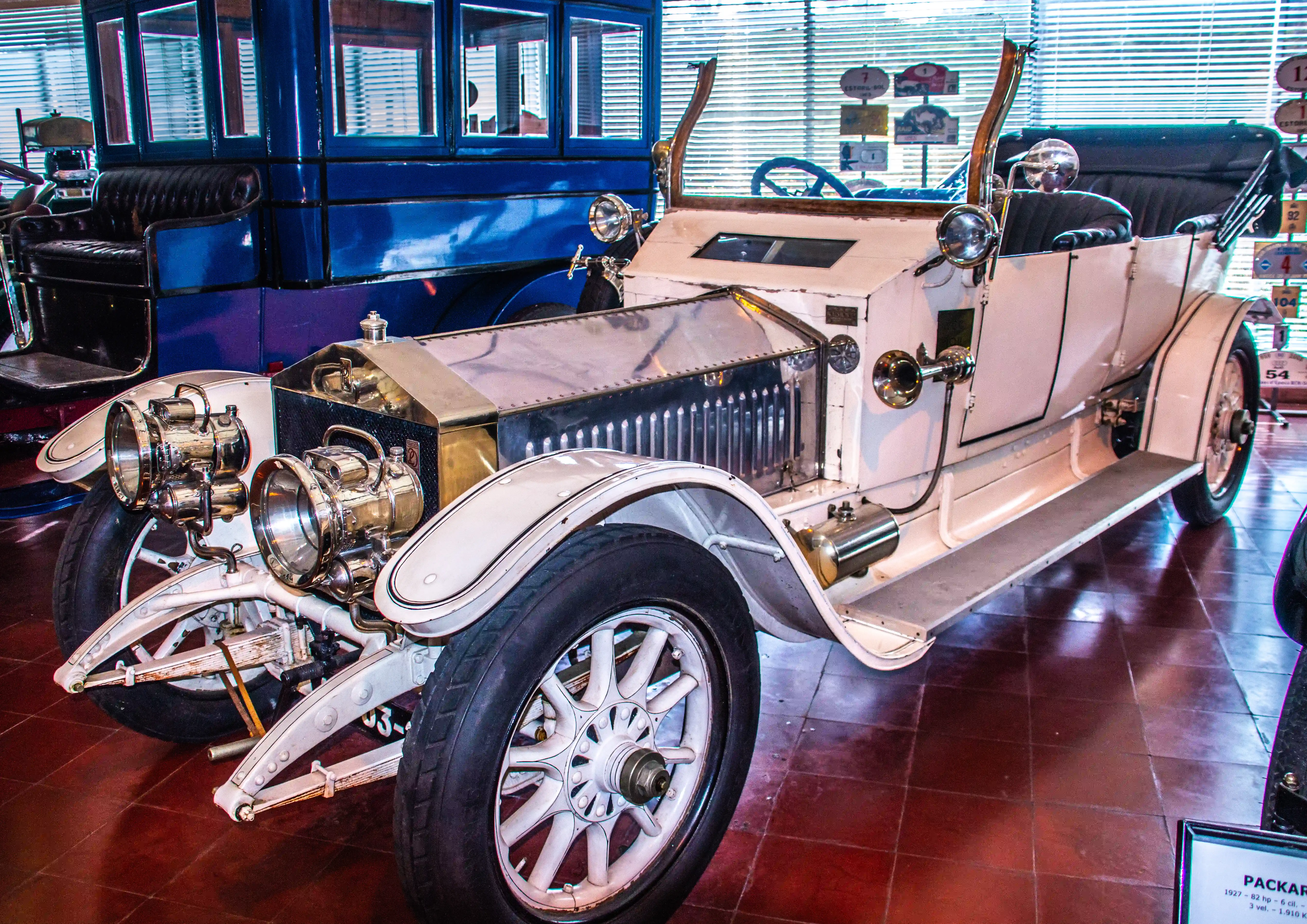 Close-up of a vintage white car with large brass headlights and white spoked wheels, parked next to a blue vintage vehicle.