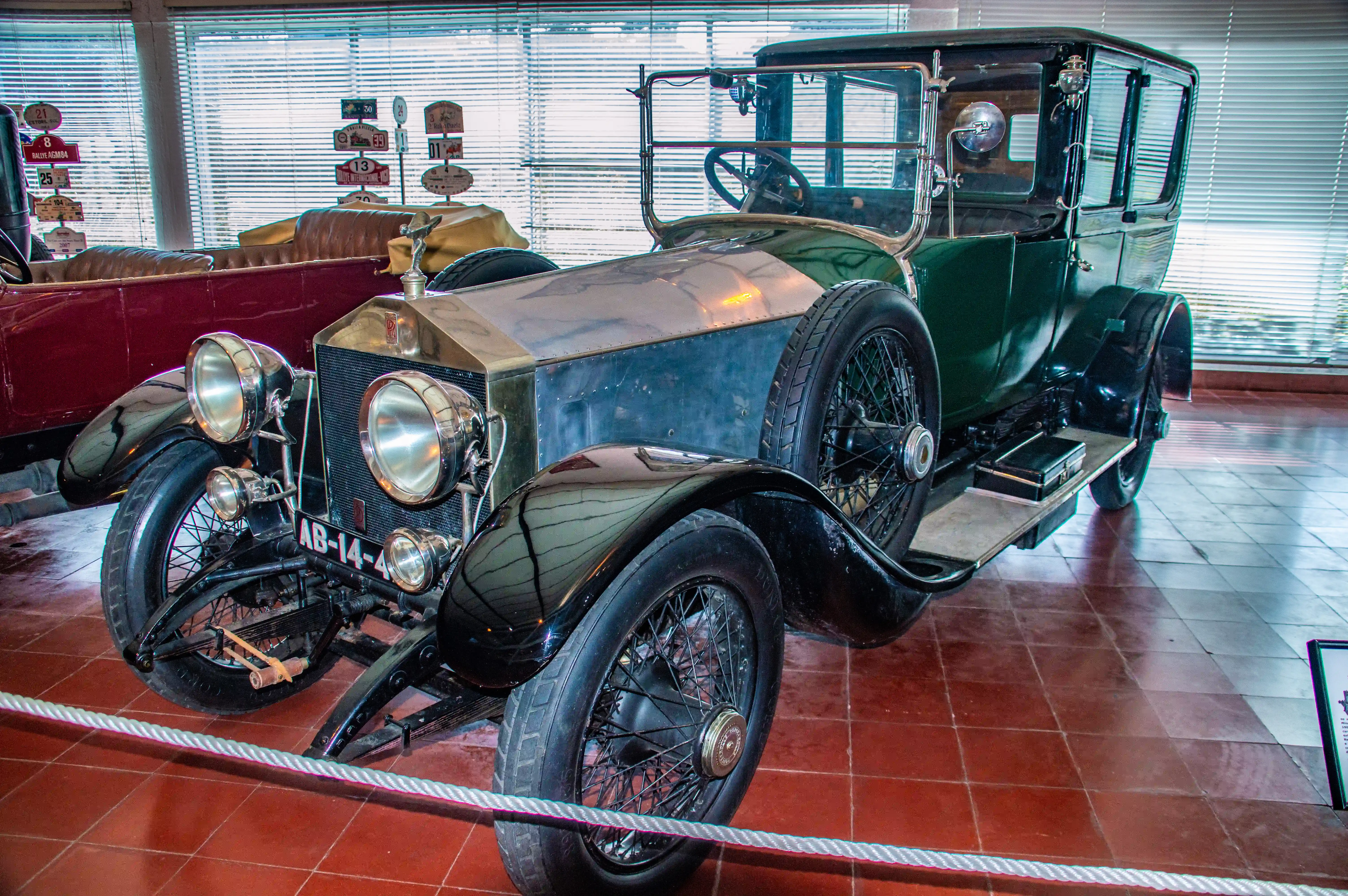 A vintage green Rolls-Royce automobile with a silver bonnet mascot and spoked wheels, displayed indoors on a red tiled floor.