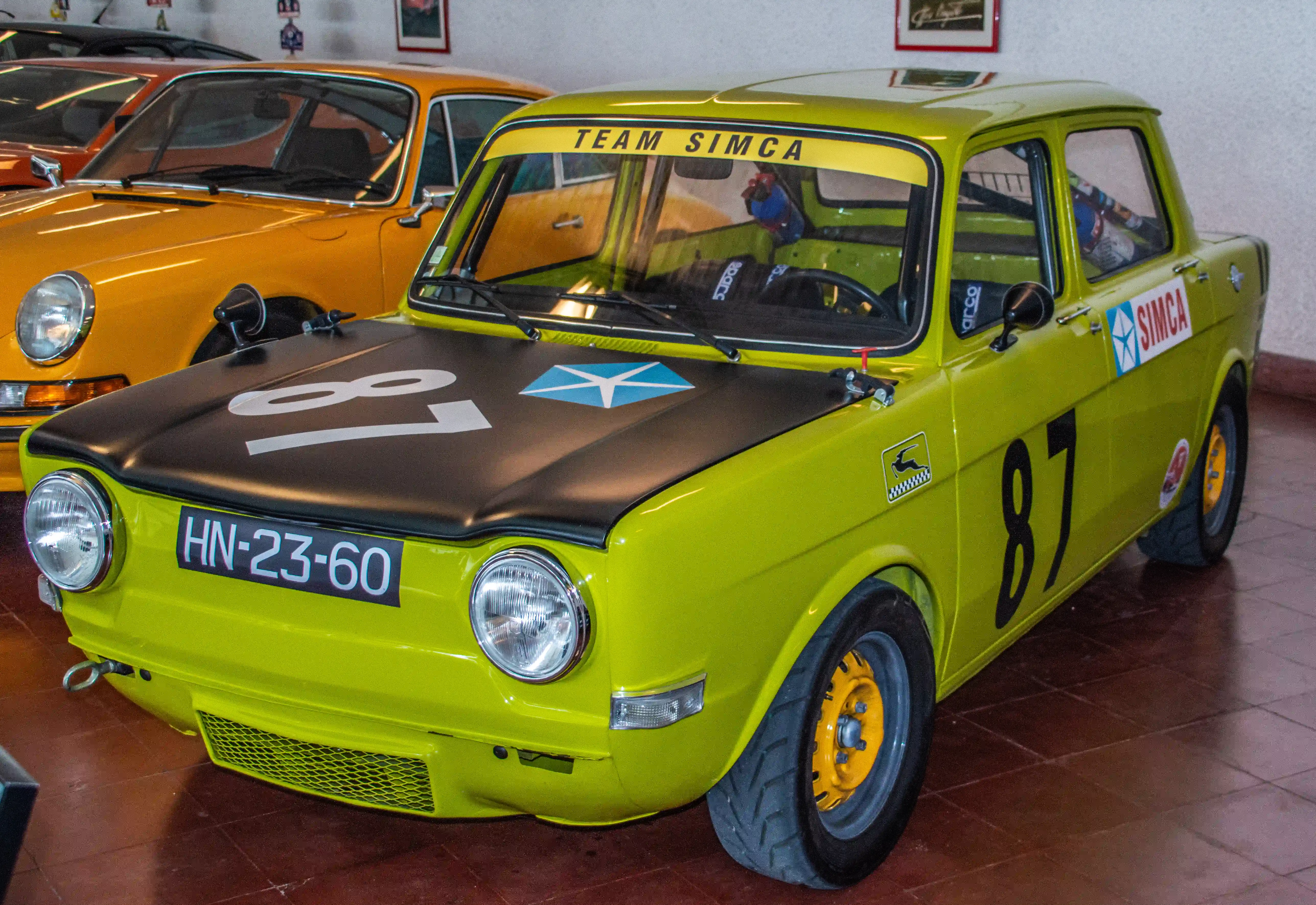 Close-up of a bright green vintage Simca rally car with the number 87 in black. The car has a black bonnet with a blue star emblem and white number 87, a yellow windscreen banner reading 