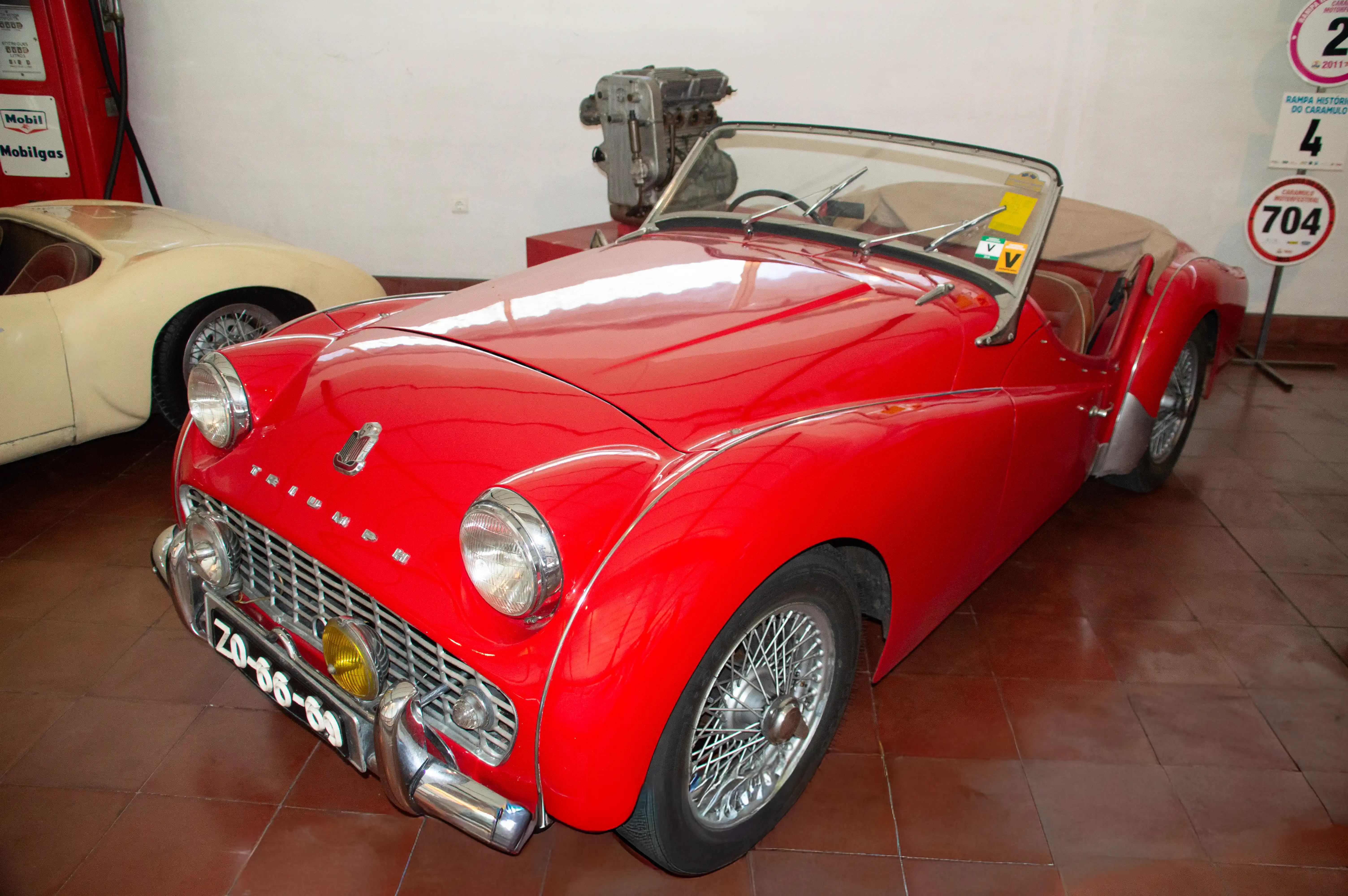 A vibrant red vintage Triumph TR3 convertible sports car is showcased indoors, possibly in a museum or classic car exhibition. The car features classic wire-spoke wheels and a beige soft-top roof. In the background, another vintage car in a cream colour is partially visible, along with a vintage Mobilgas fuel pump. An engine unit is also displayed behind the Triumph. Signage indicating a historical event or competition is visible on the wall to the right.
