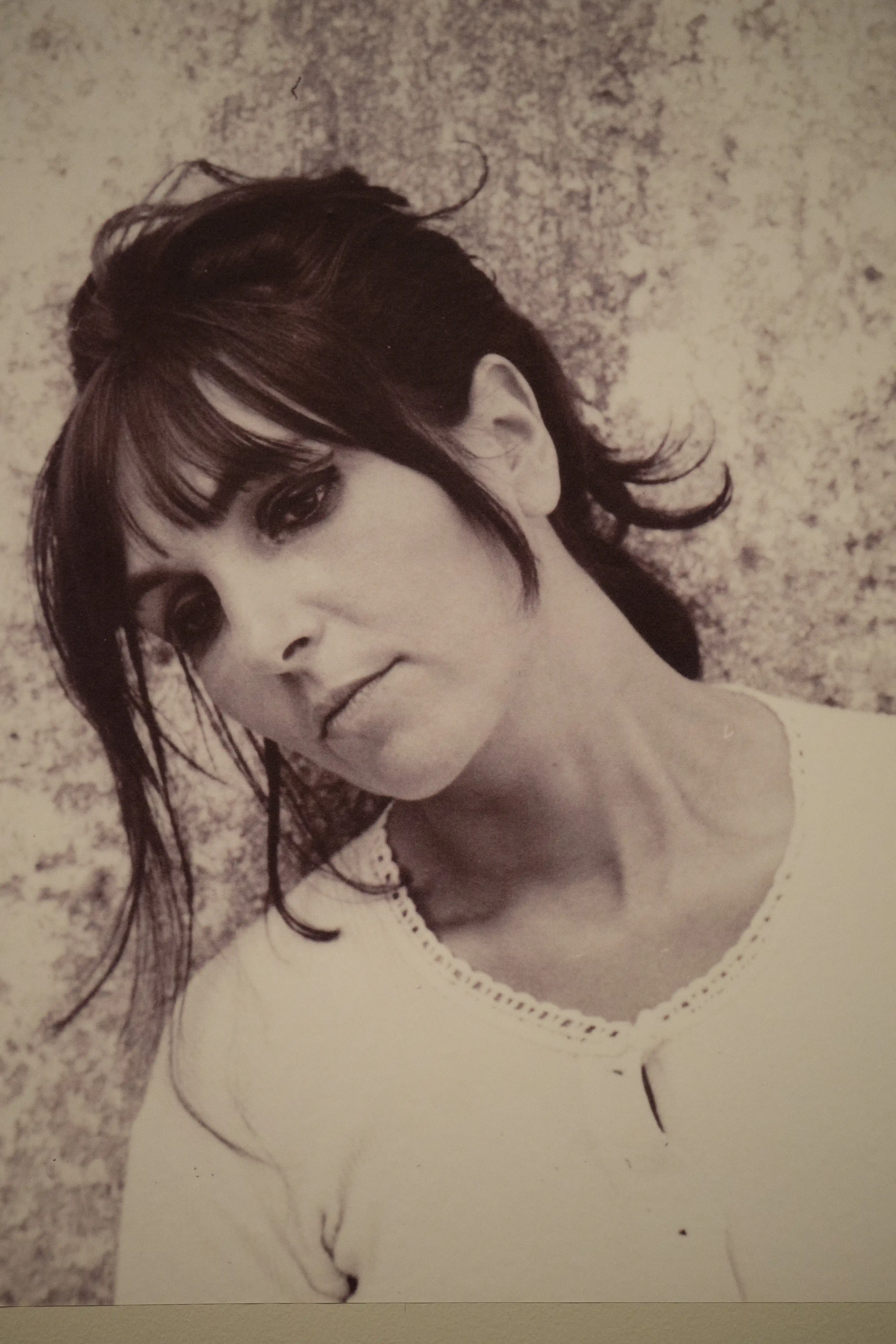  Black and white close-up portrait of a woman with dark, tousled hair, bangs, and heavy eyeliner, looking thoughtfully at the camera. She wears a white top with an embroidered neckline.