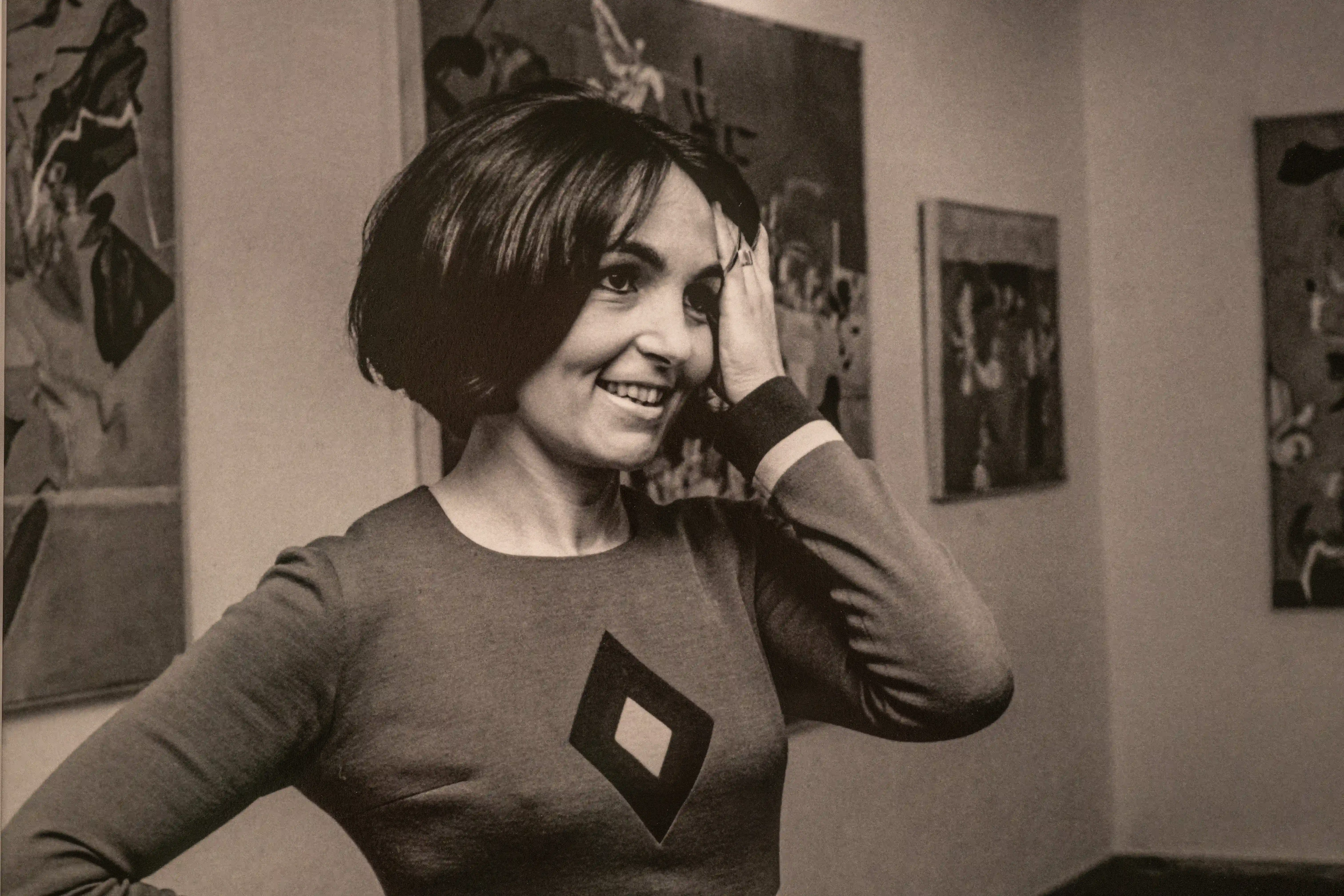 A black and white photograph showing a Paulo Rego with short dark hair smiling and holding her hand to her forehead. She is wearing a dark dress with a diamond shape on the front. There are paintings on the wall behind her.