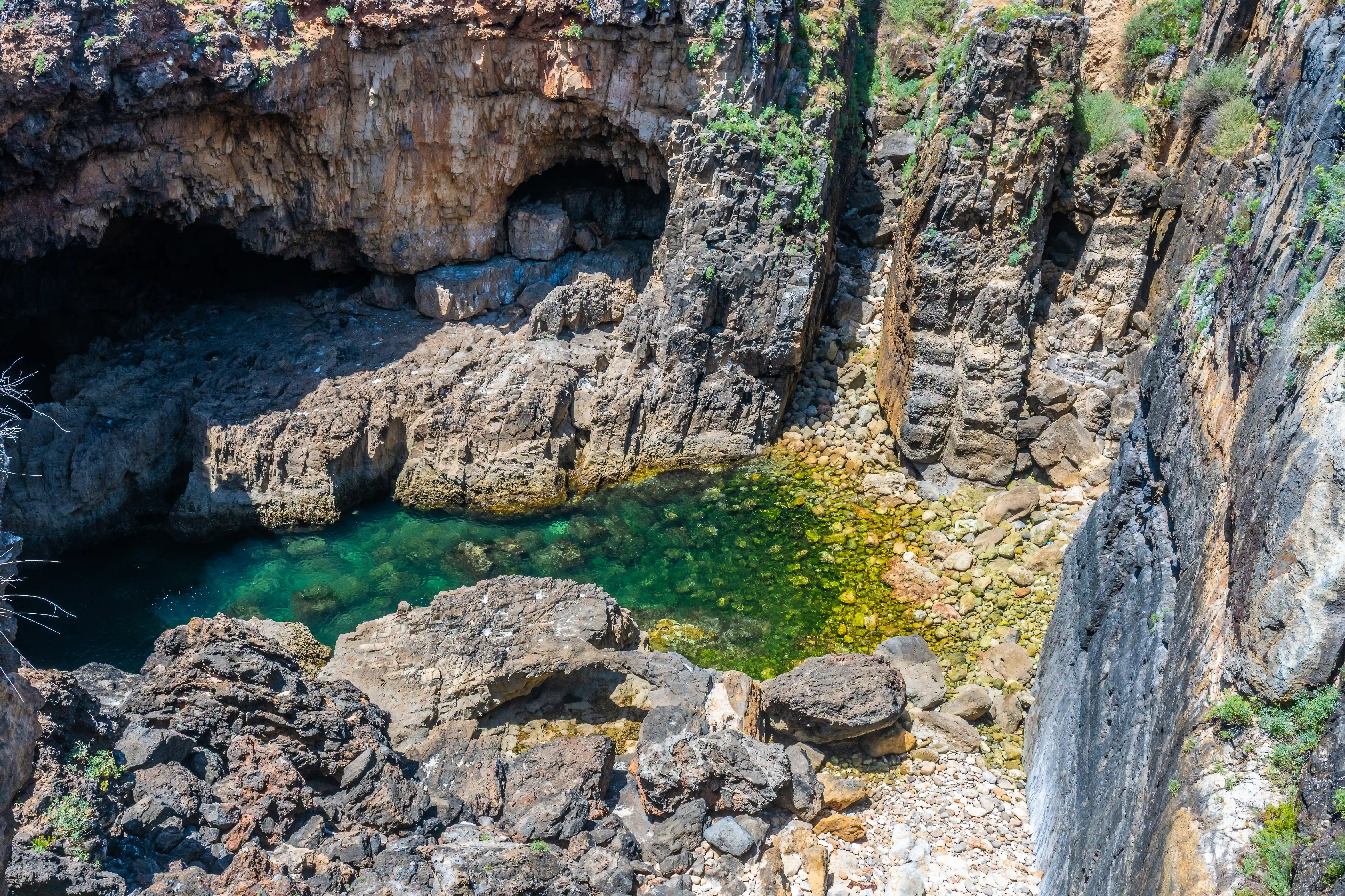 A high-angle shot of a secluded cove featuring a dark cave opening into rugged, layered rock formations. A small body of clear, green water is visible in the centre, surrounded by a rocky shore and cliffs.