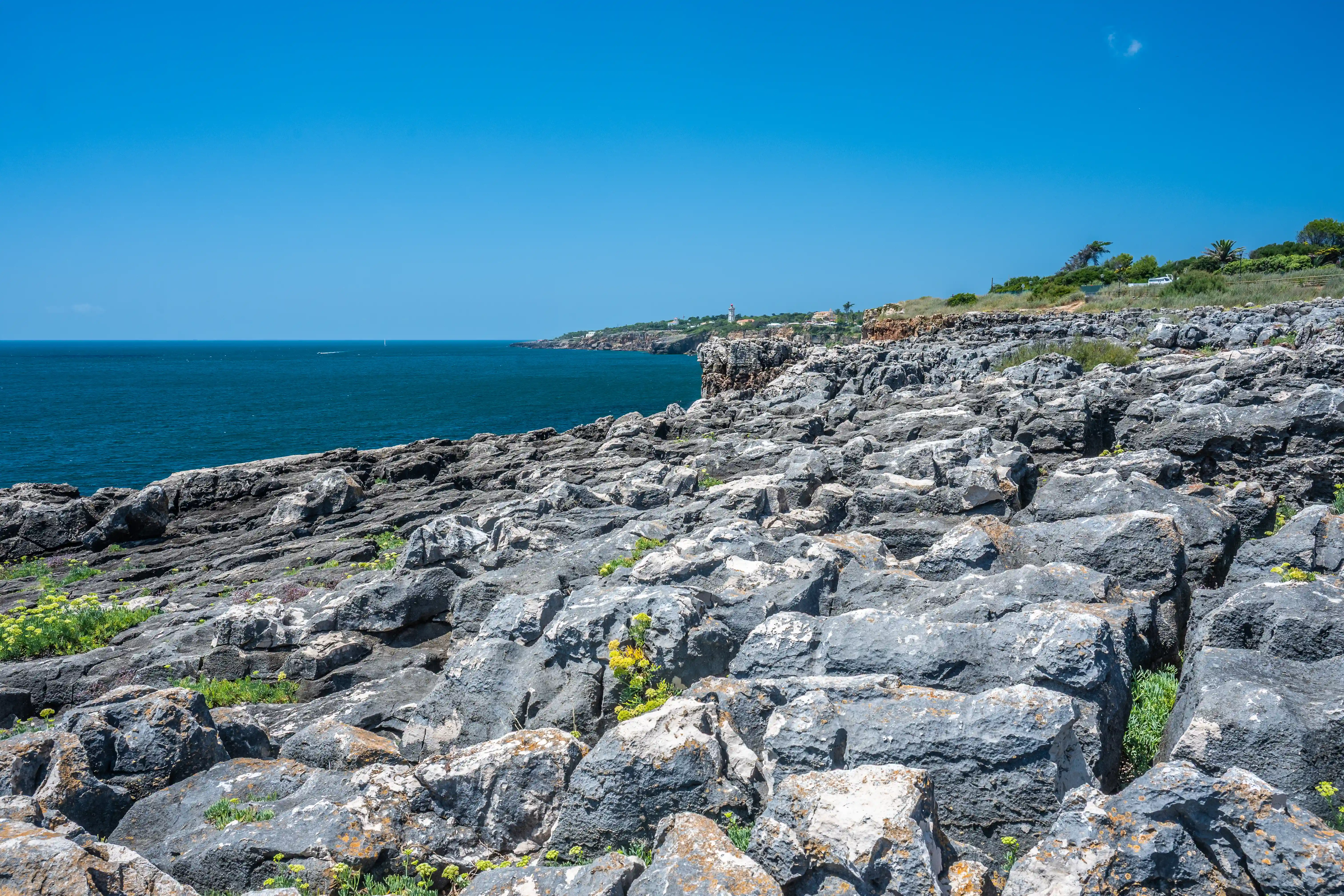 A wide shot of a rocky coastline. The foreground is filled with large, grey, weathered rocks, some with patches of green vegetation. The deep blue sea stretches to the horizon on the left, meeting a hazy coastline in the distance with a few buildings and a lighthouse visible. The sky is a clear, bright blue.