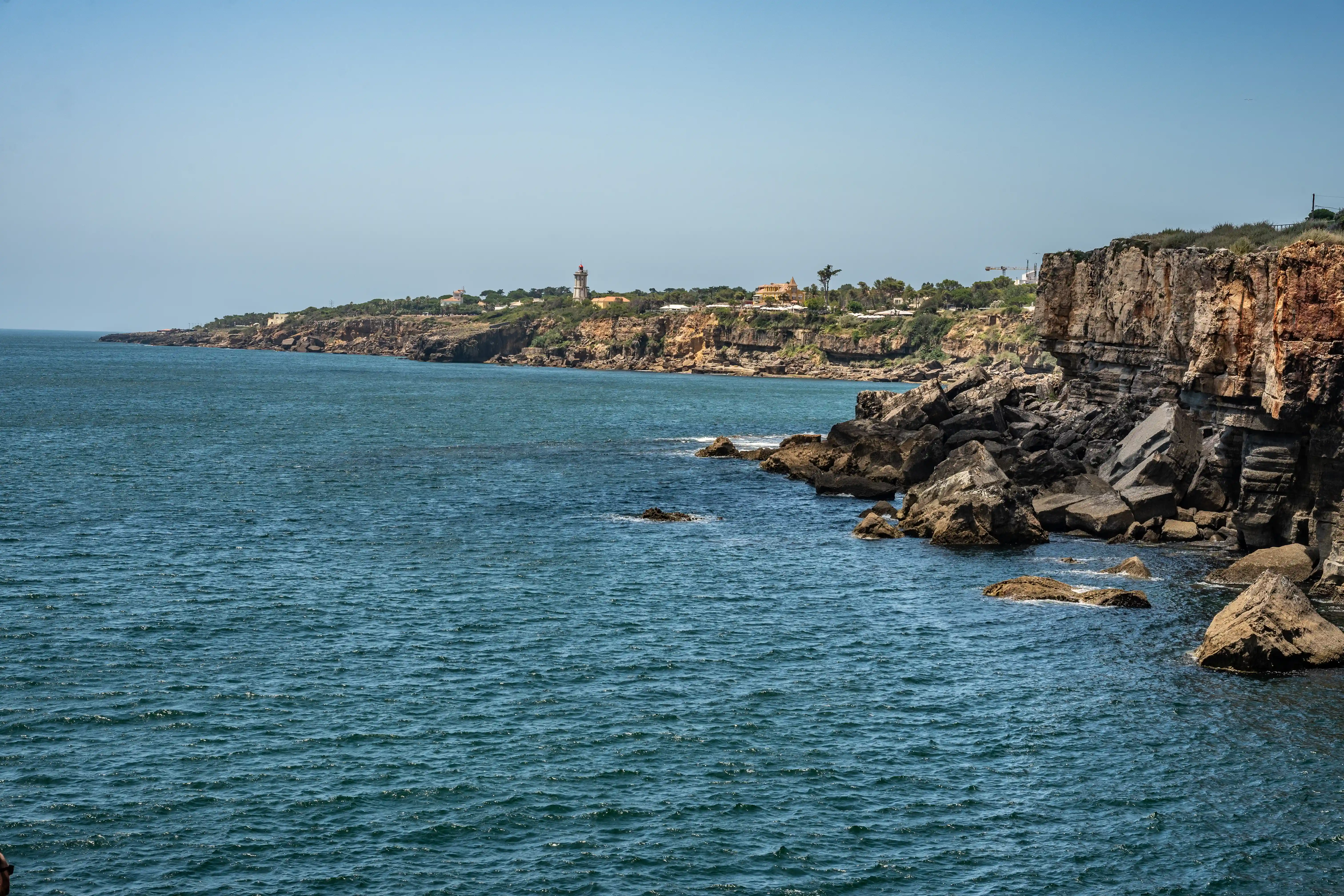 Wide shot of a rugged, rocky coastline meeting the blue ocean under a clear sky. A lighthouse and buildings are visible on the clifftop in the distance.