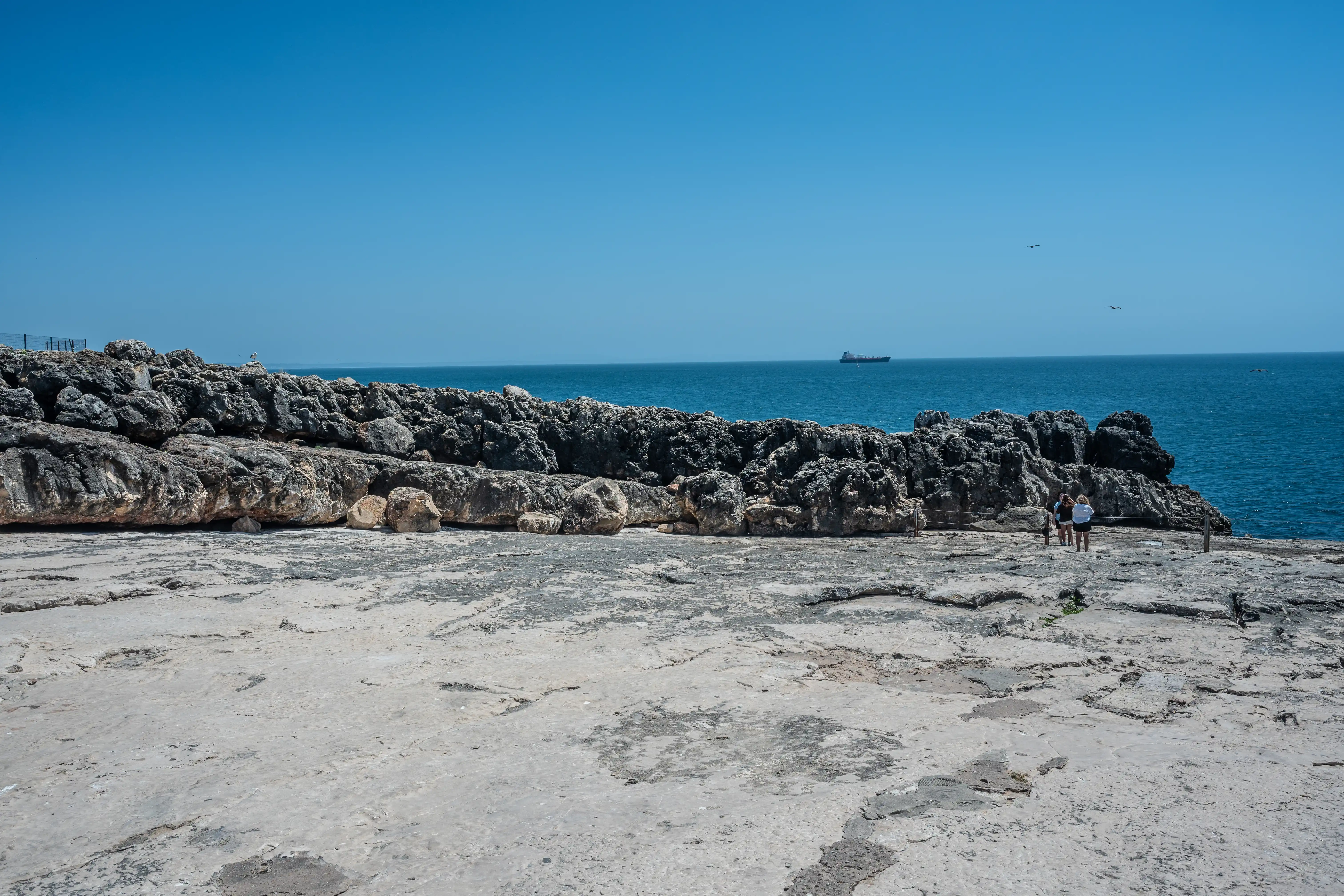 A wide shot of a textured, light-coloured rocky terrain leading to a dark, jagged rock formation along a coastline. Two figures are visible standing on the rocks. In the distance, the deep blue sea meets the clear sky, with a cargo ship sailing on the horizon and two small birds in the air.