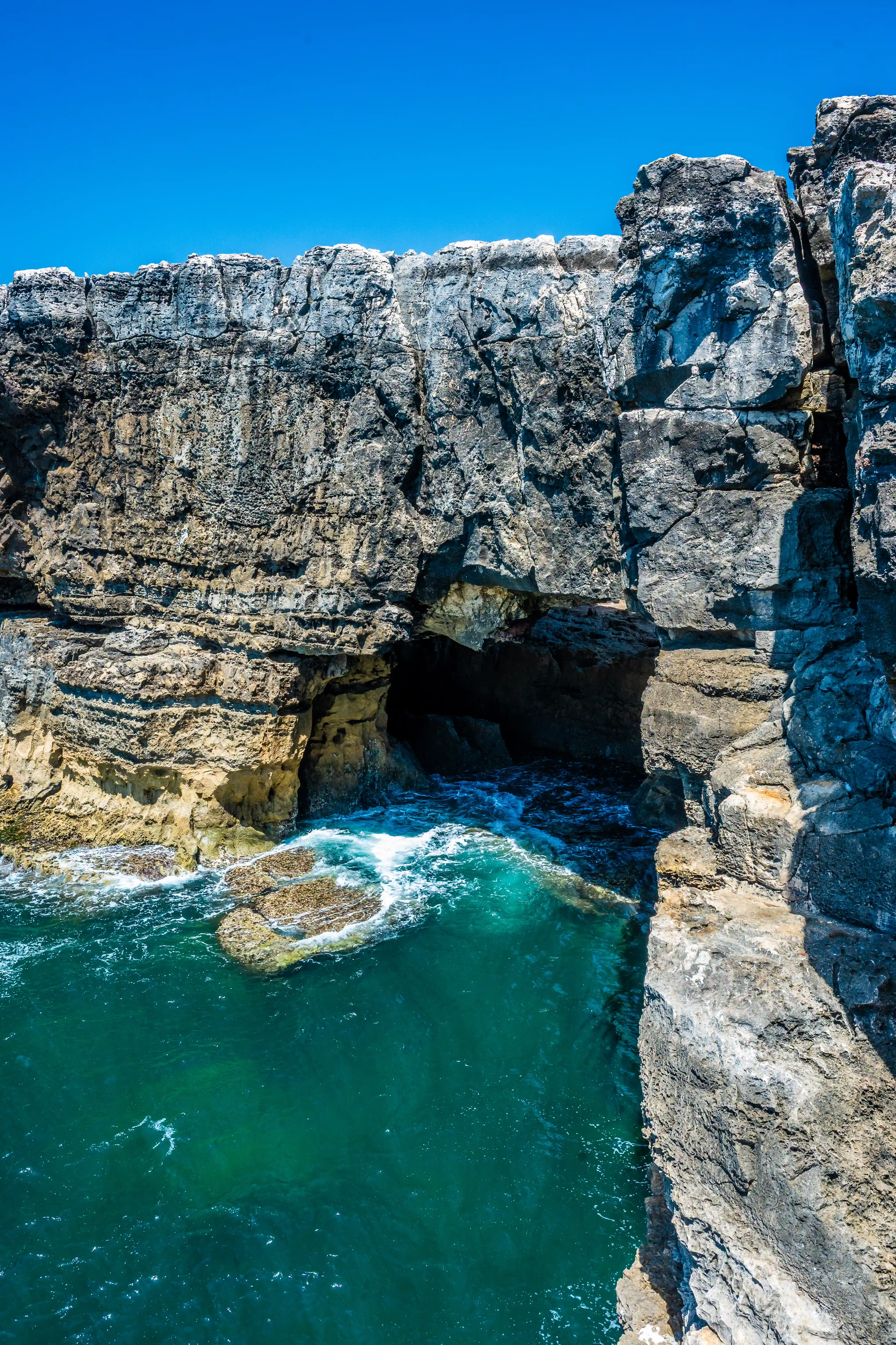 A view looking down into a cave carved into a steep, grey cliff face. Turquoise water with white waves laps against rocks at the entrance to the cave, under a bright blue sky.