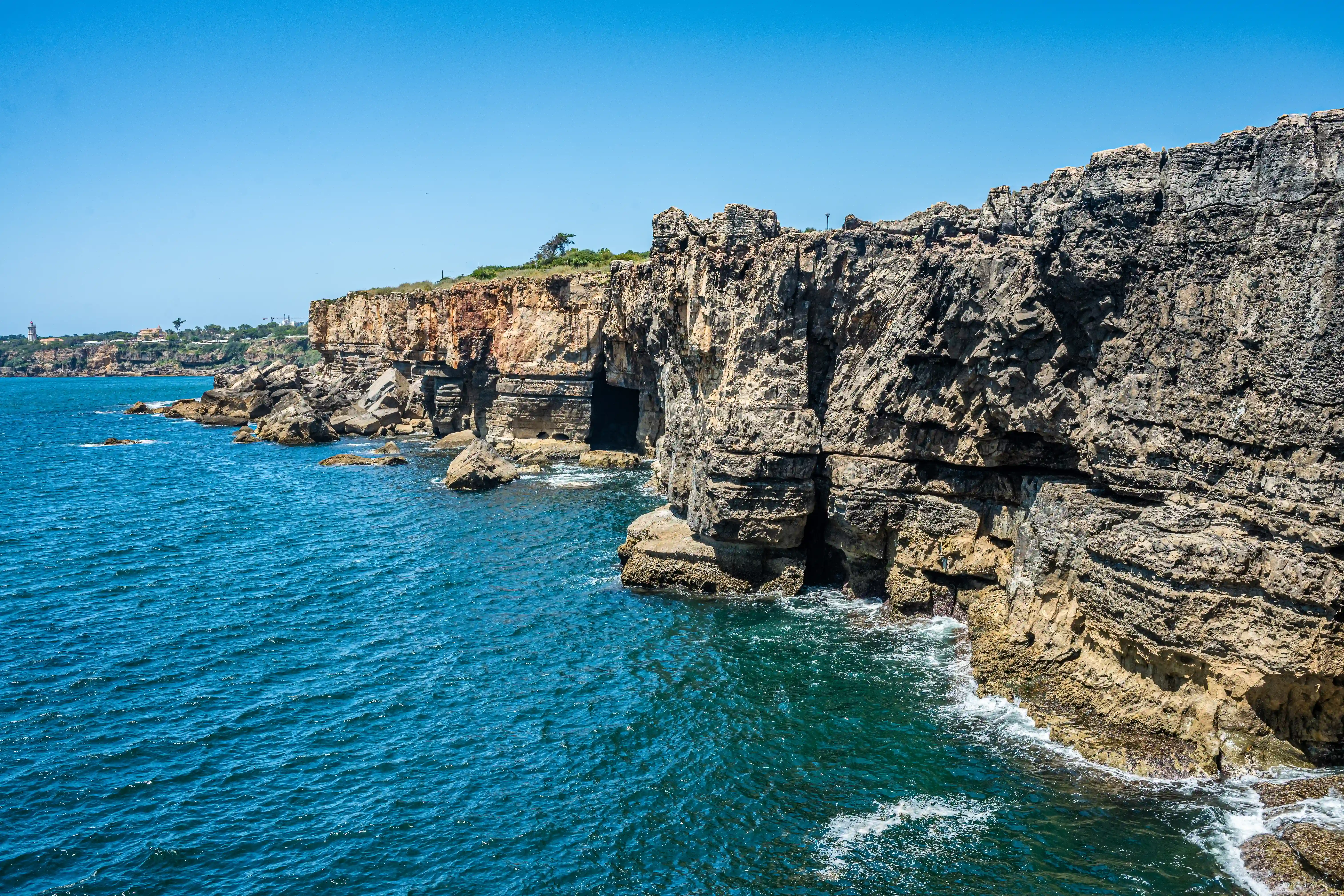 A panoramic view of imposing, layered coastal cliffs meeting a deep blue, rippling sea under a cloudless sky. Waves crash against the rocks at the base of the cliffs, revealing natural caves and rough rock textures.