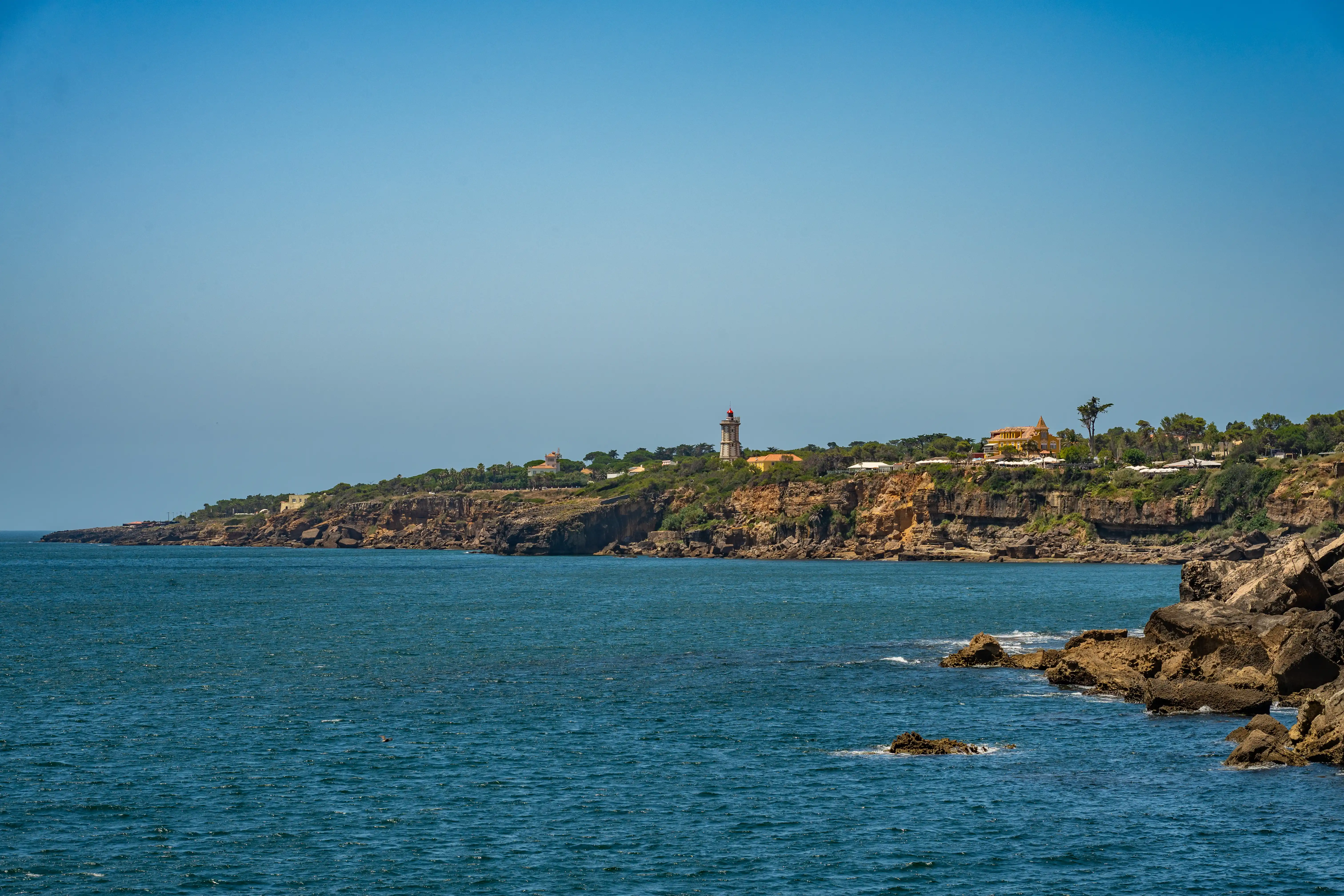  A wide shot of a cliffside overlooking the ocean. A lighthouse stands on the cliff, with other buildings and trees dotting the landscape. The sea is dark blue, and the sky is a pale blue.