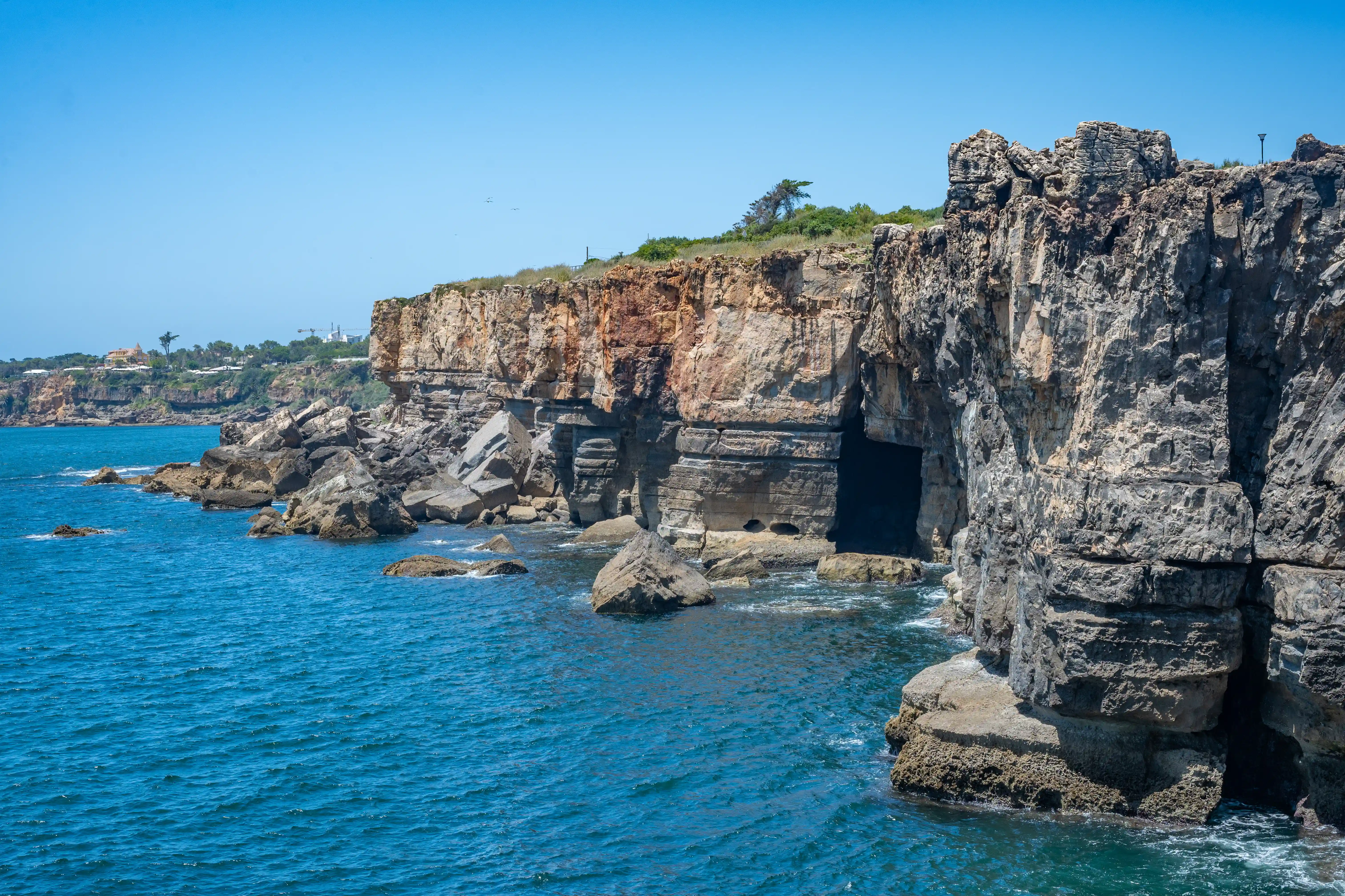 A view of imposing coastal cliffs with visible sea caves and scattered rocks in the foreground, lapped by the blue sea.