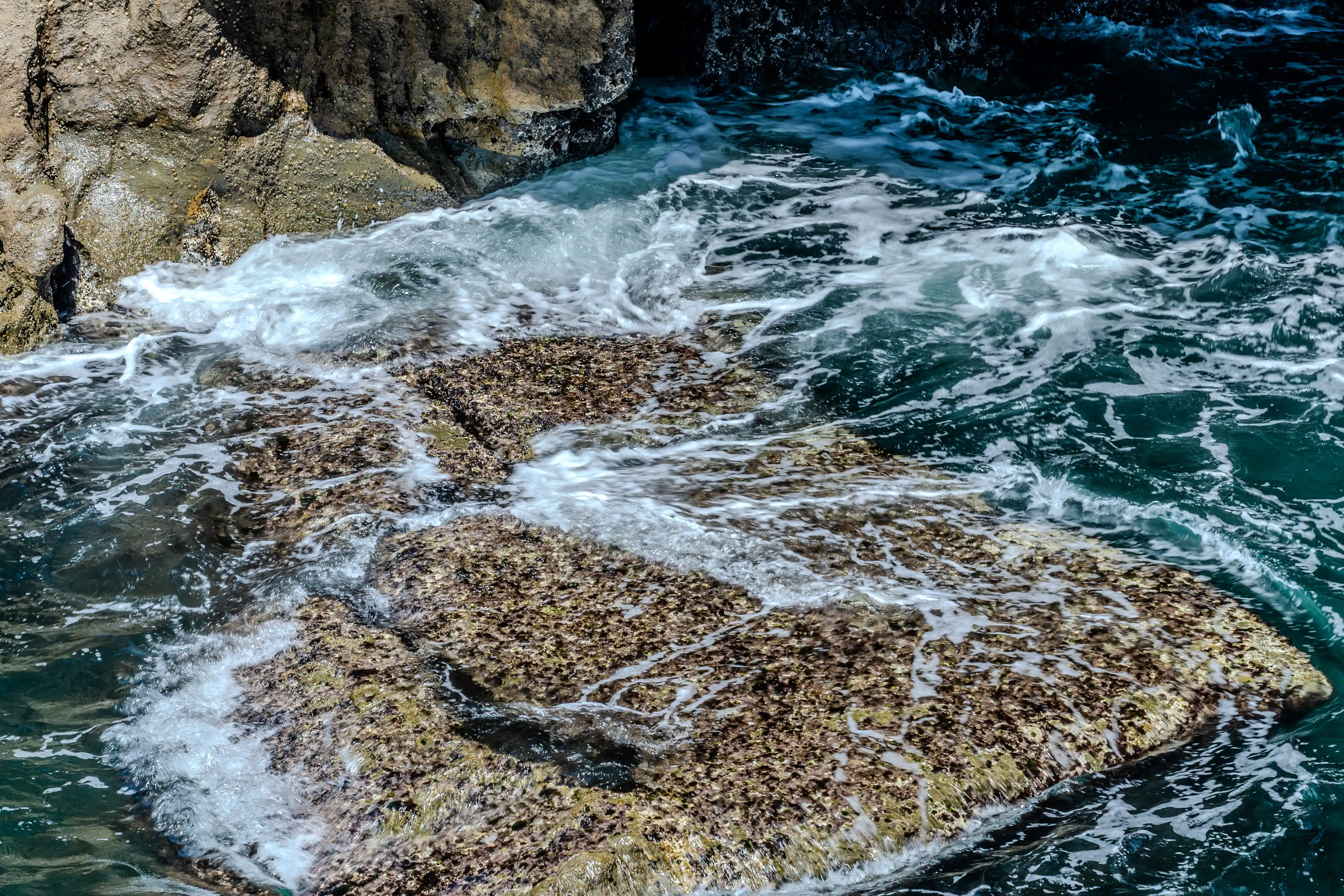 A high-angle view of dark blue water swirling around rugged, barnacle-covered rocks, with white sea foam frothing around their edges.