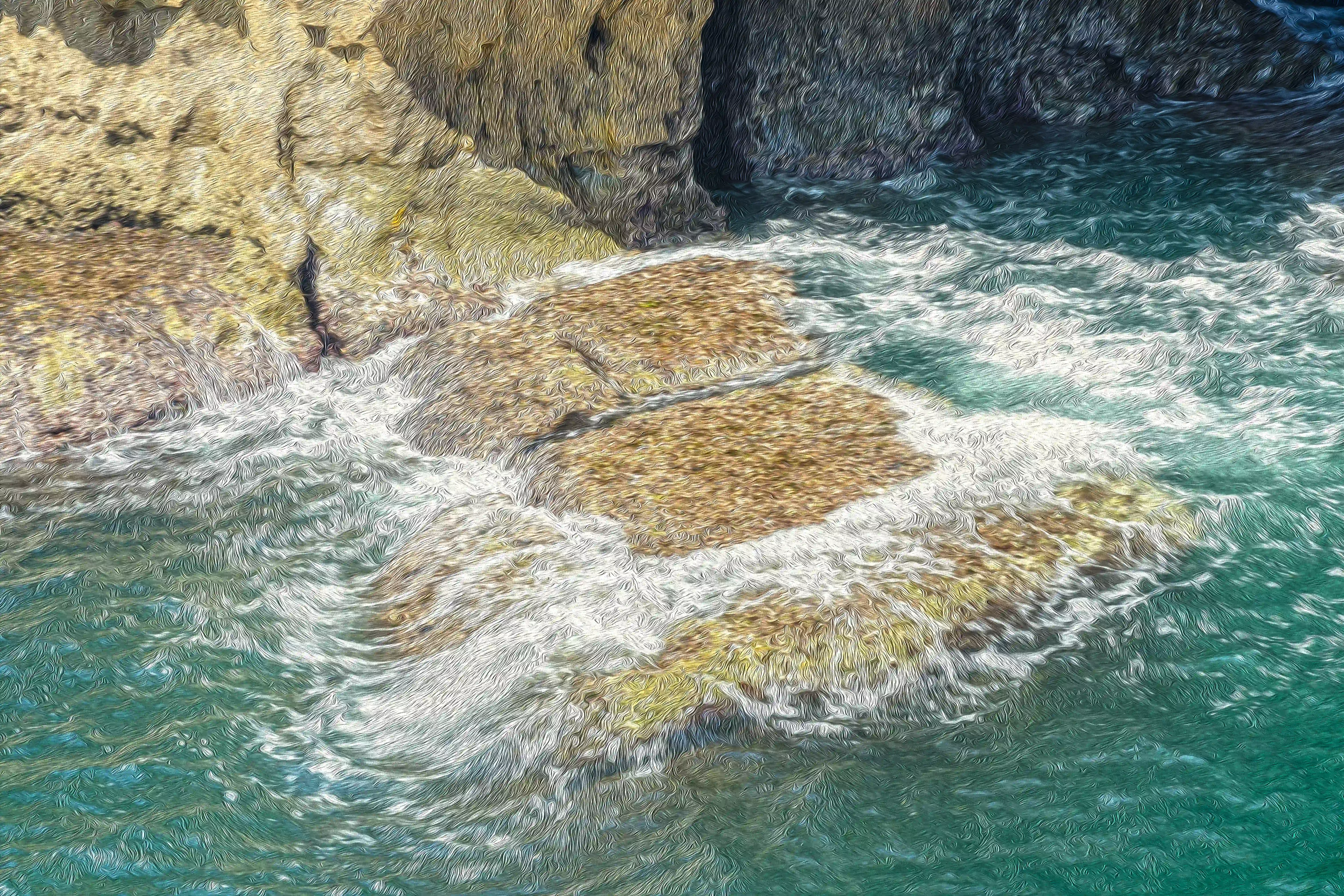  A high-angle view of choppy, dark turquoise sea water swirling around and over a flat, textured rock formation that is partially submerged. Sunlight illuminates parts of the rock and the white foam of the waves. In the background, dark, shadowed cliffs rise up.