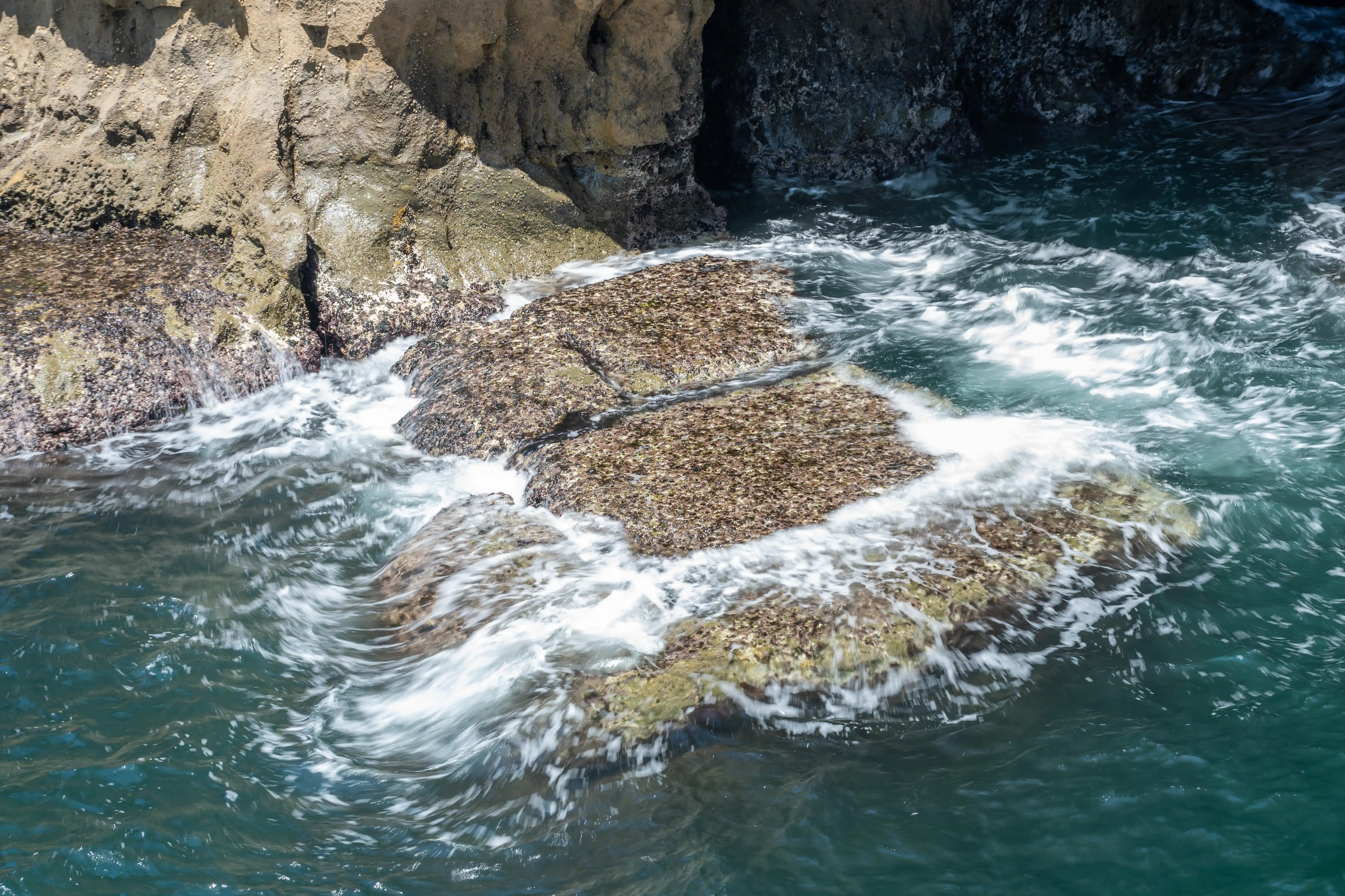  A high-angle view of choppy, dark turquoise sea water swirling around and over a flat, textured rock formation that is partially submerged. Sunlight illuminates parts of the rock and the white foam of the waves. In the background, dark, shadowed cliffs rise up.