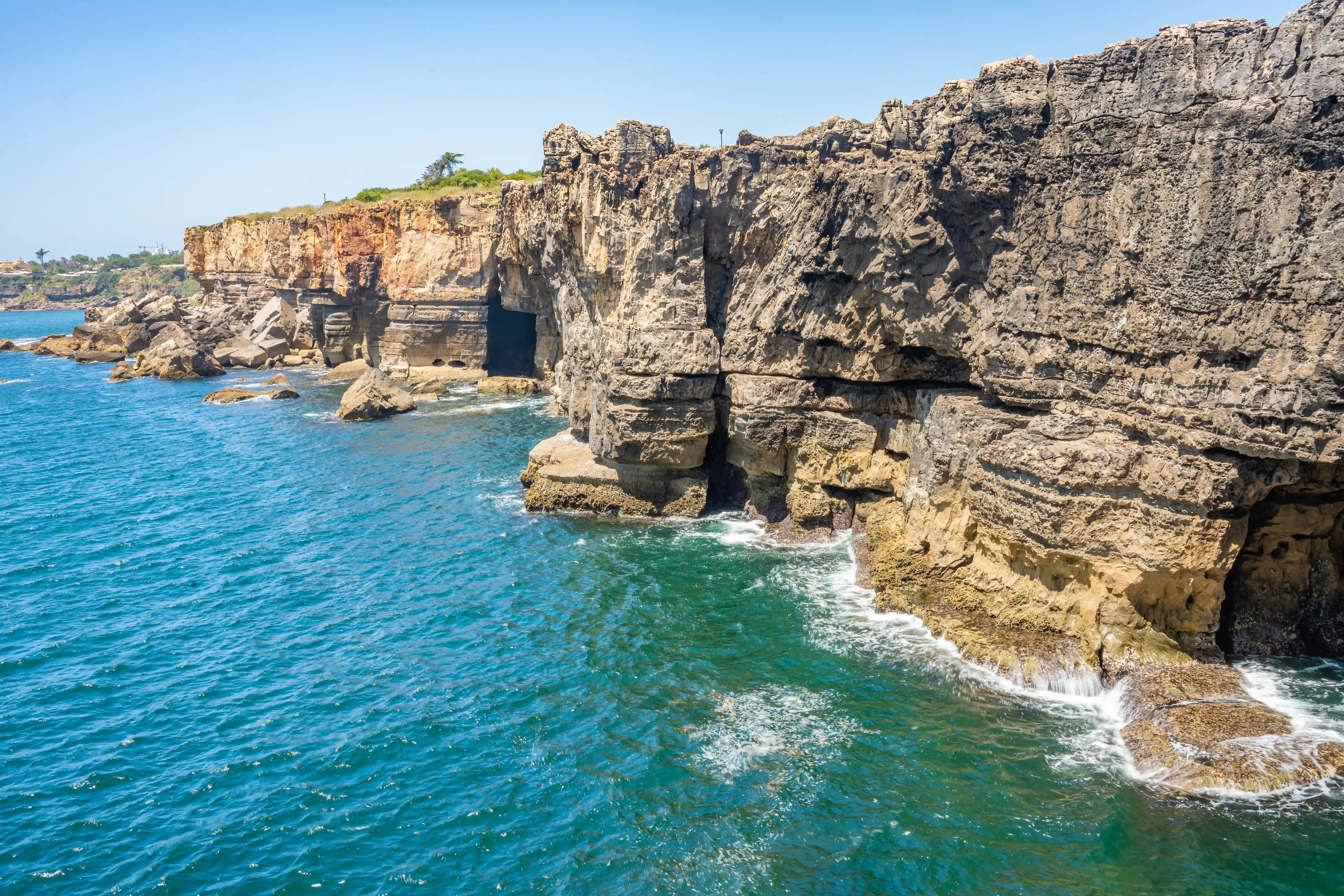 A wide shot of dramatic, layered sea cliffs with caves, meeting the deep blue ocean under a clear