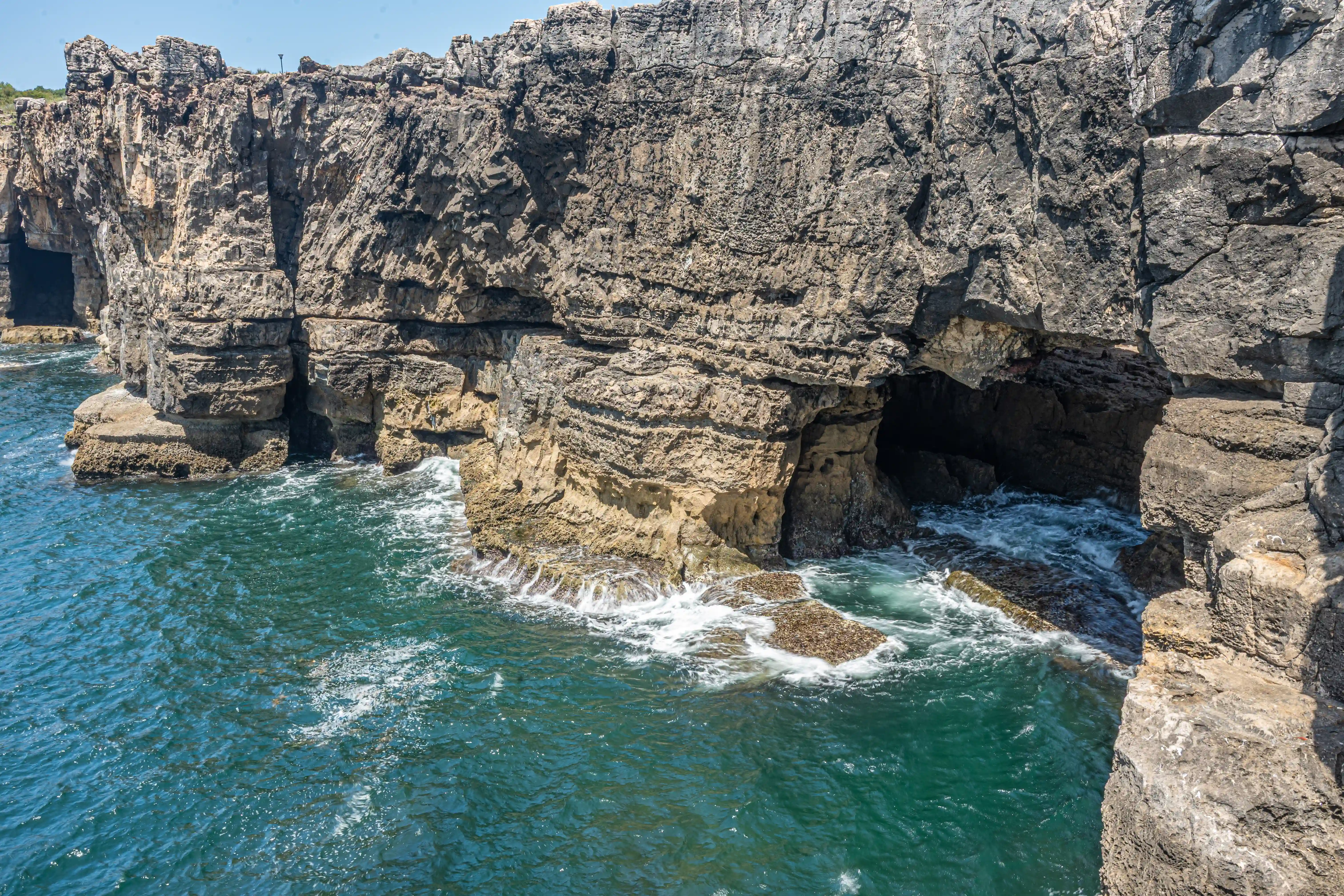 A wide shot of dramatic, layered sea cliffs with dark caves, as turquoise ocean water crashes against the rocks below.